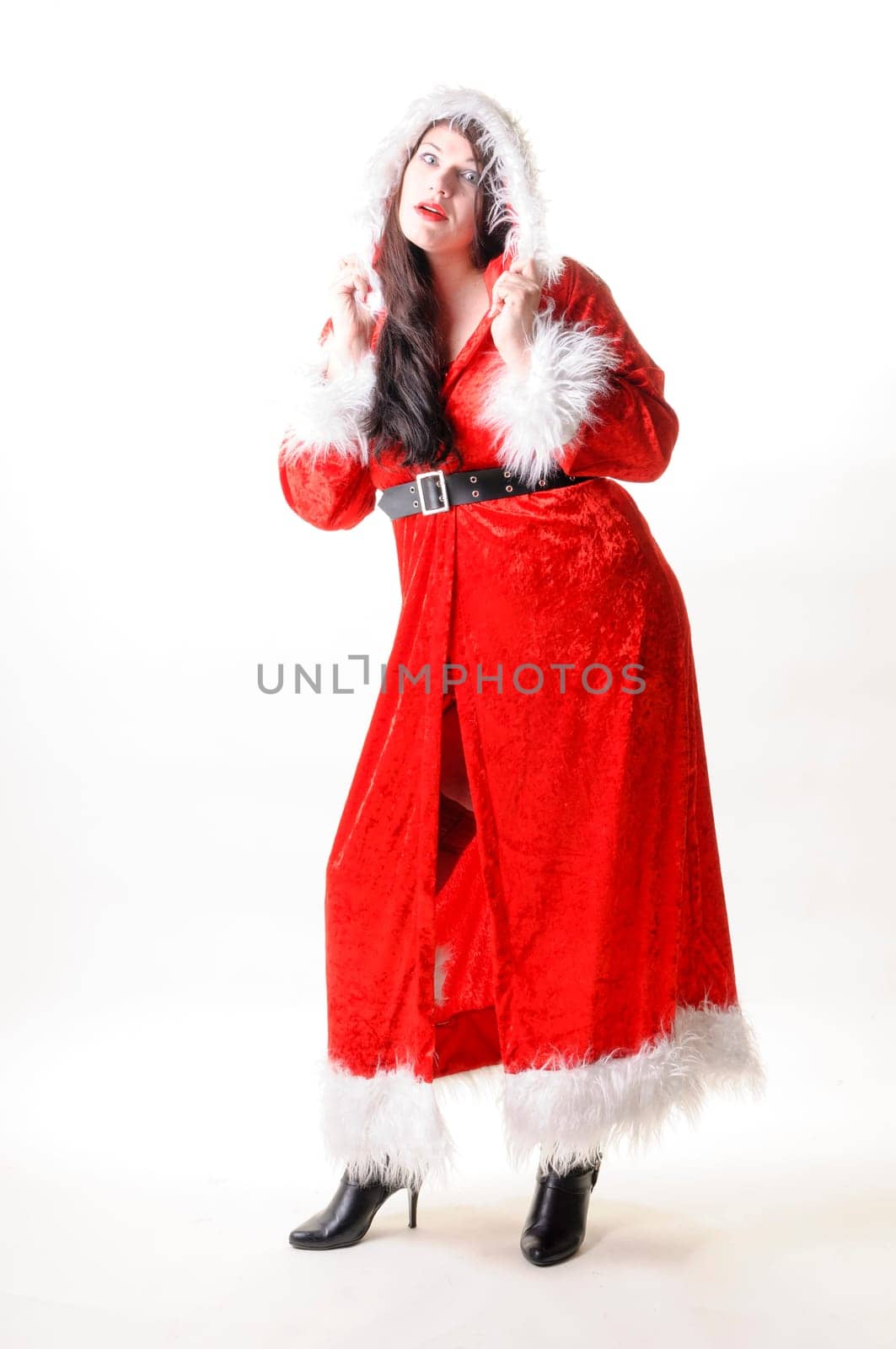 Adult overweight woman with long dark hair wears a red Christmas coat and black boots and stands free in front of a white background.