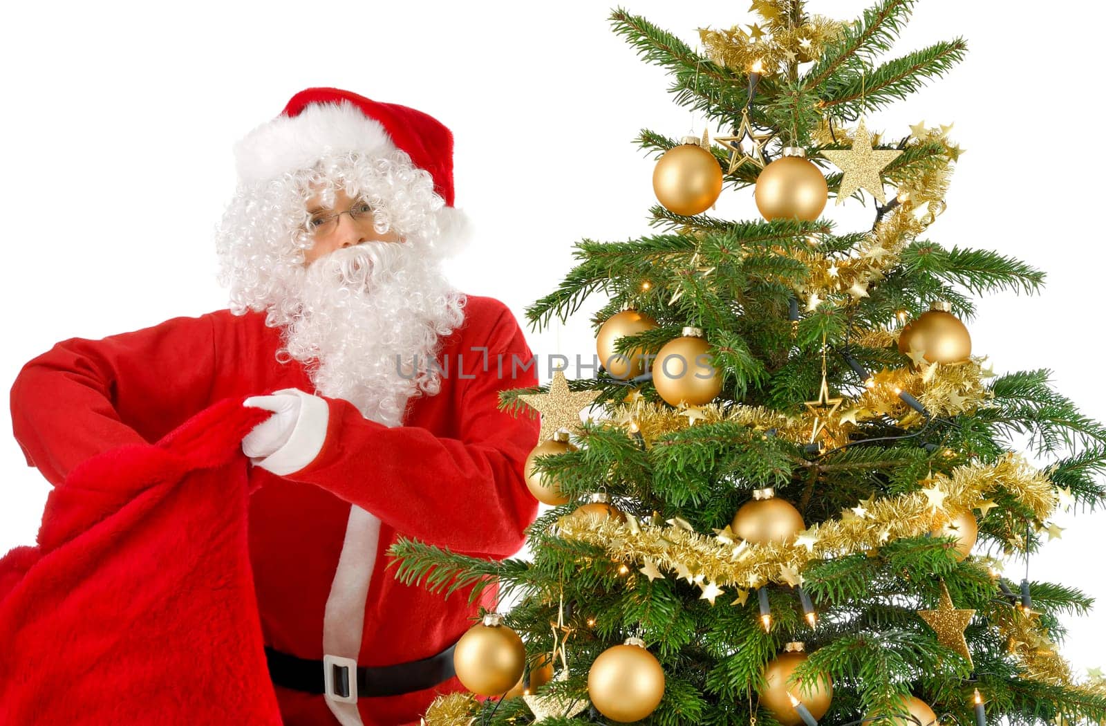 Santa Claus stands next to the Christmas tree and reaches into his red gift bag