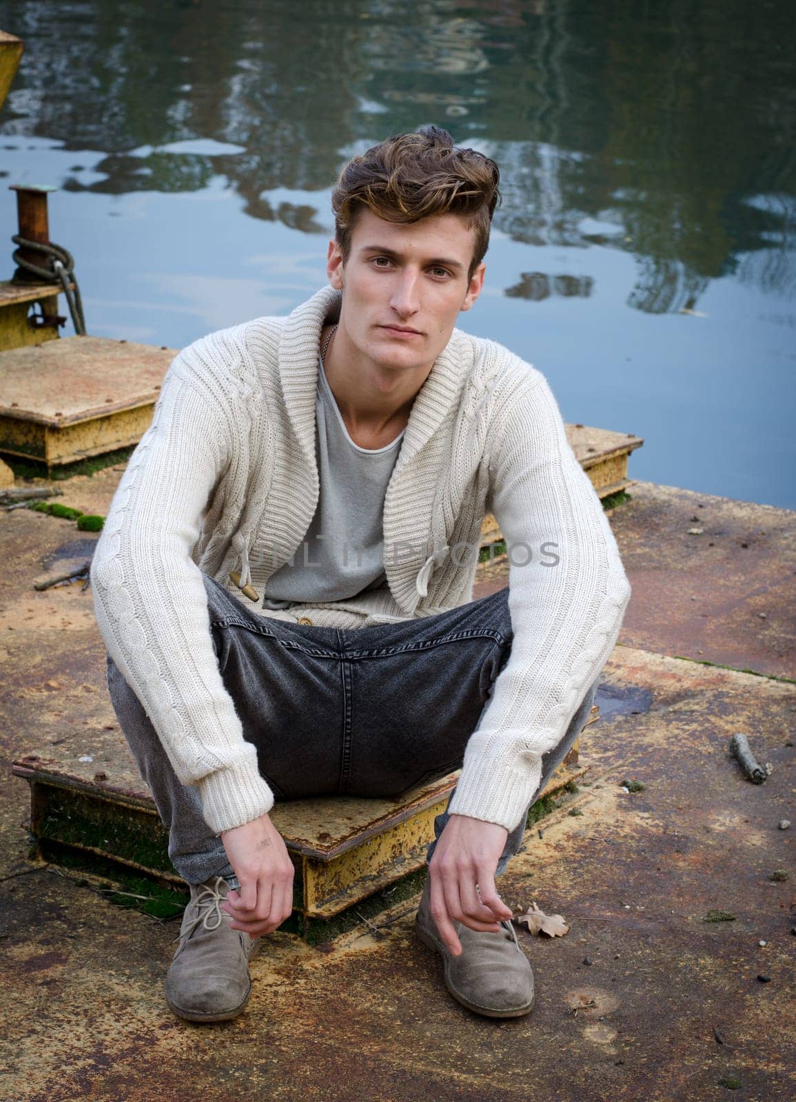 Attractive young man sitting on rusty metal next to river waters, looking at camera