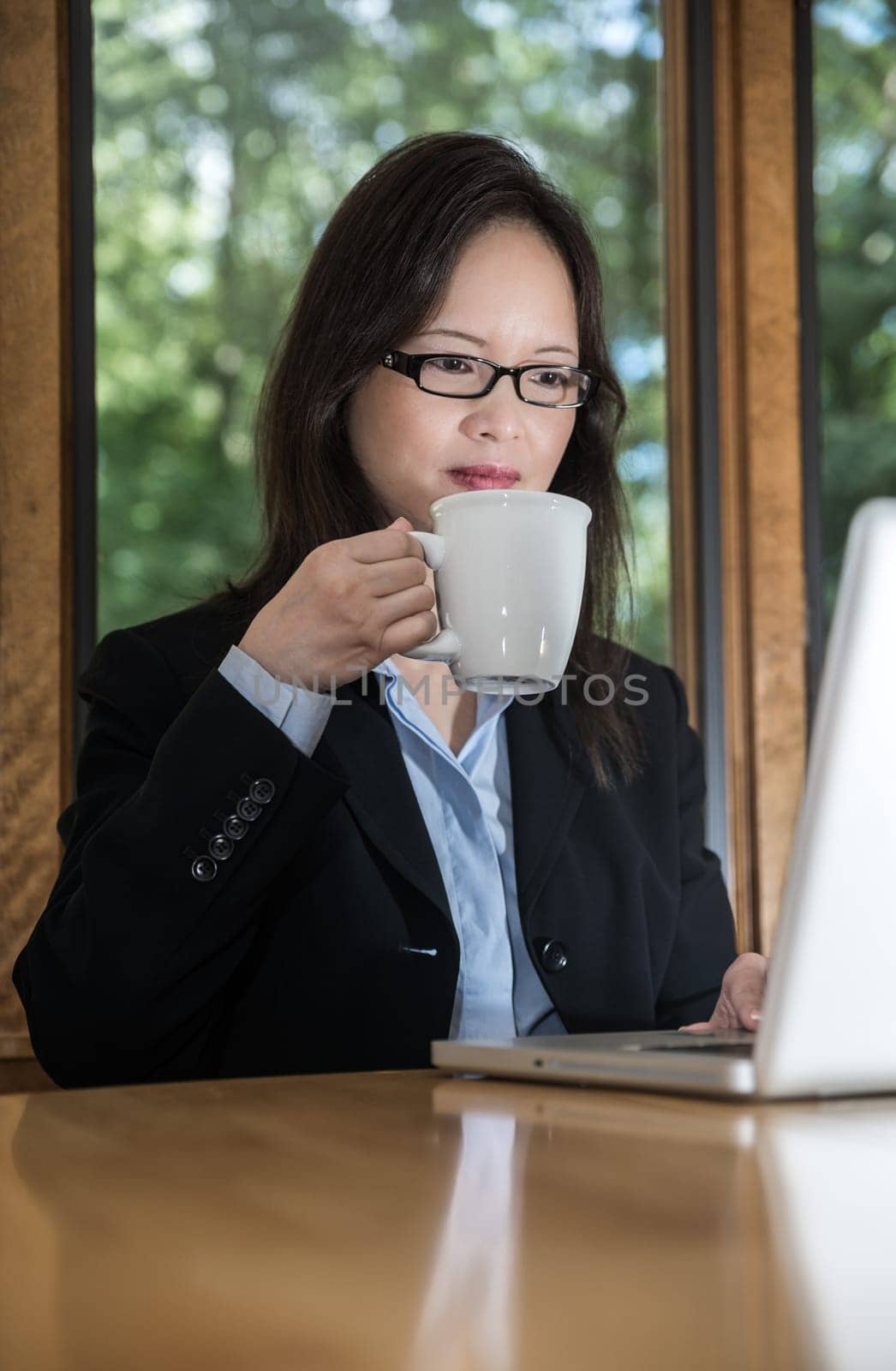 Woman in business suit with laptop on desk and drinking coffee in front of a window