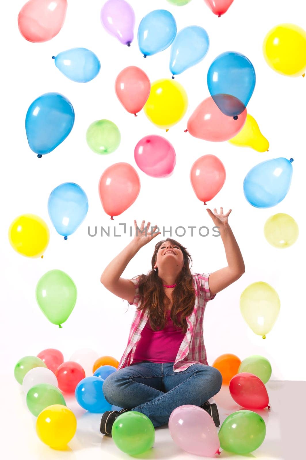 Teenage girl sits on the floor and stretches her hands up to catch colorful balloons against a white background