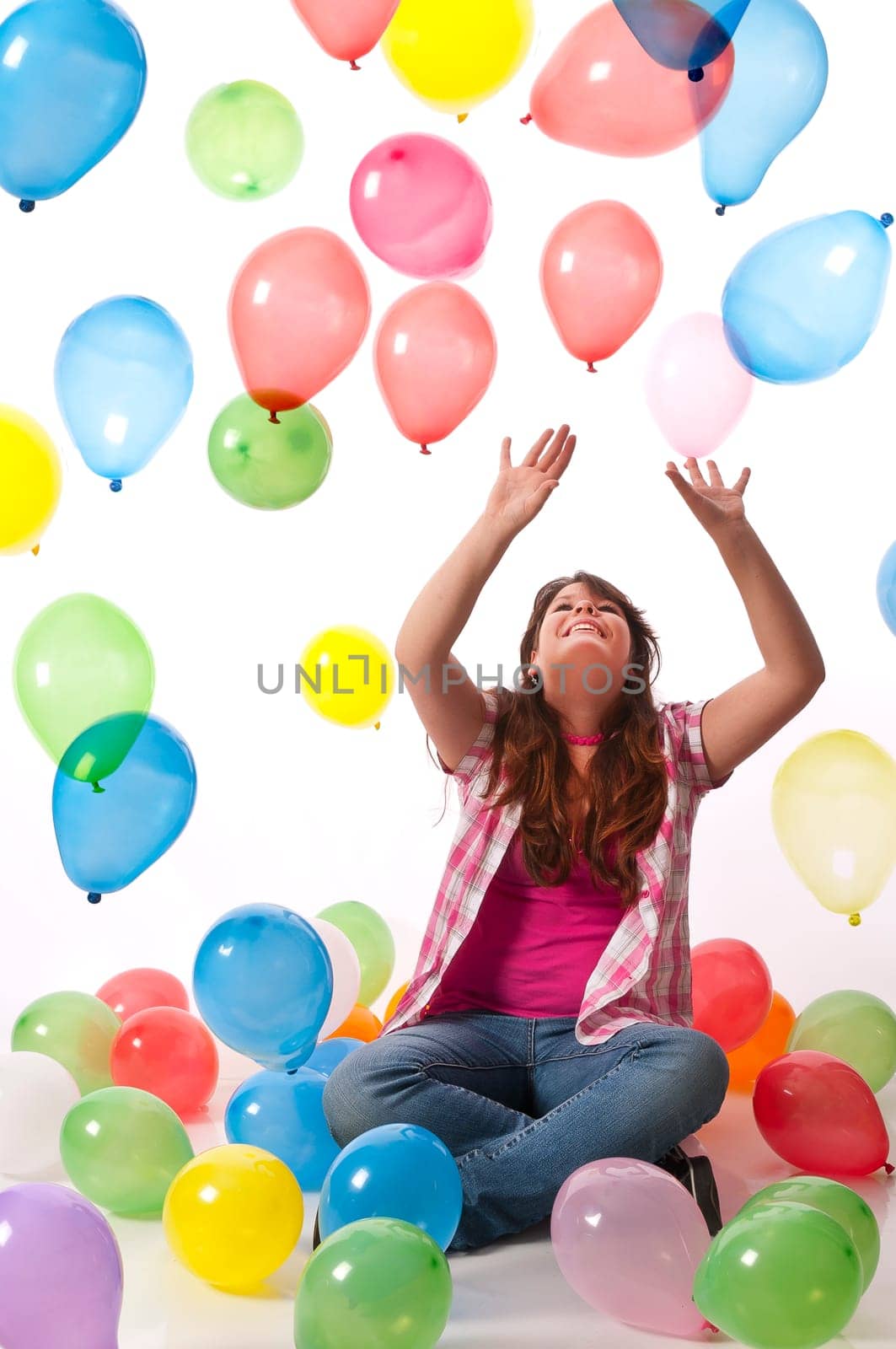 Teenage girl sits on the floor and stretches her hands up to catch colorful balloons against a white background
