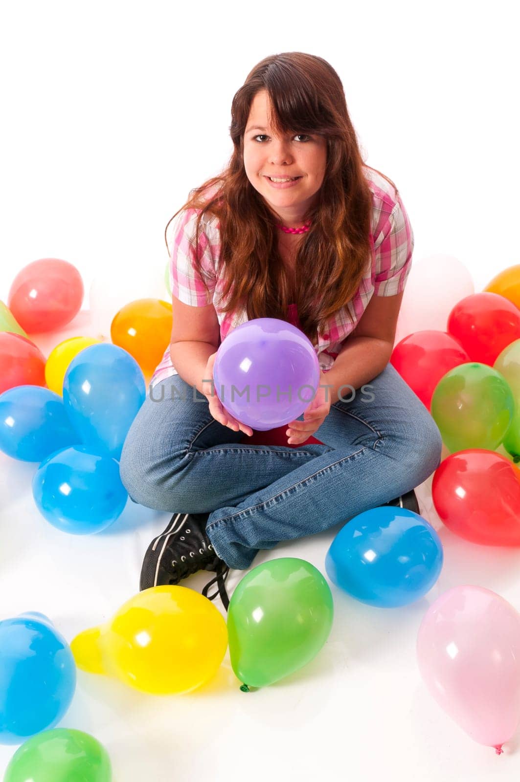 Teenage girl sits on the floor and stretches her hands up to catch colorful balloons against a white background