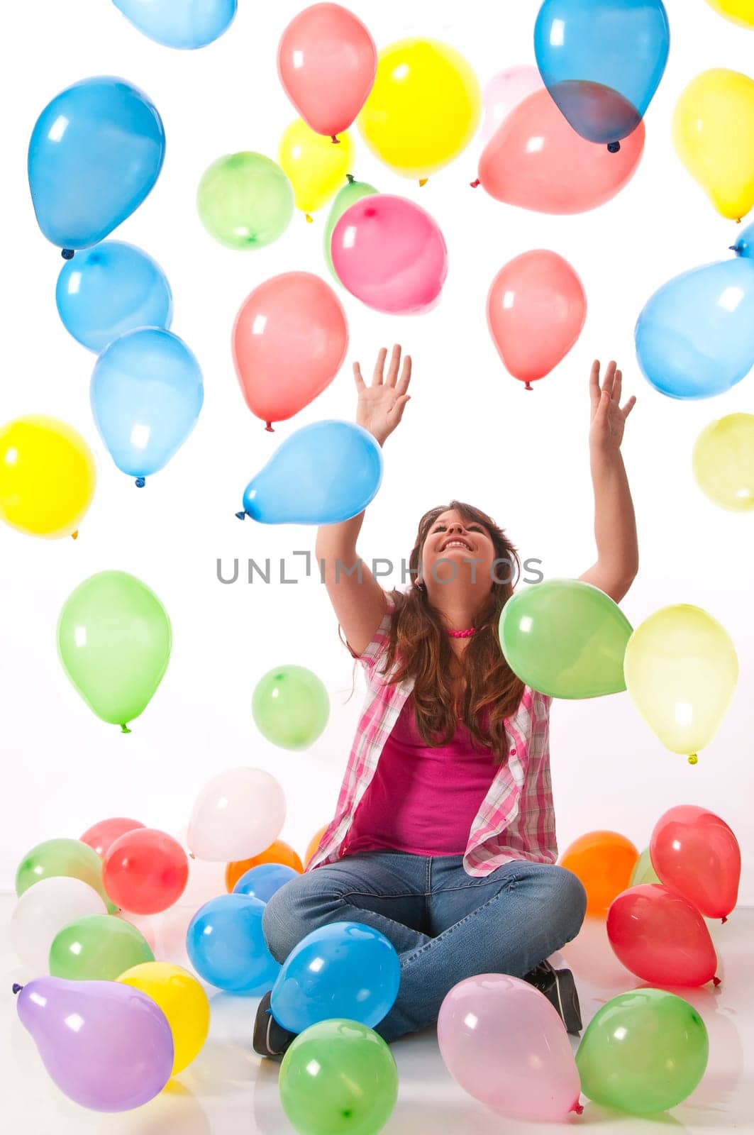 Teenage girl sits on the floor and stretches her hands up to catch colorful balloons against a white background