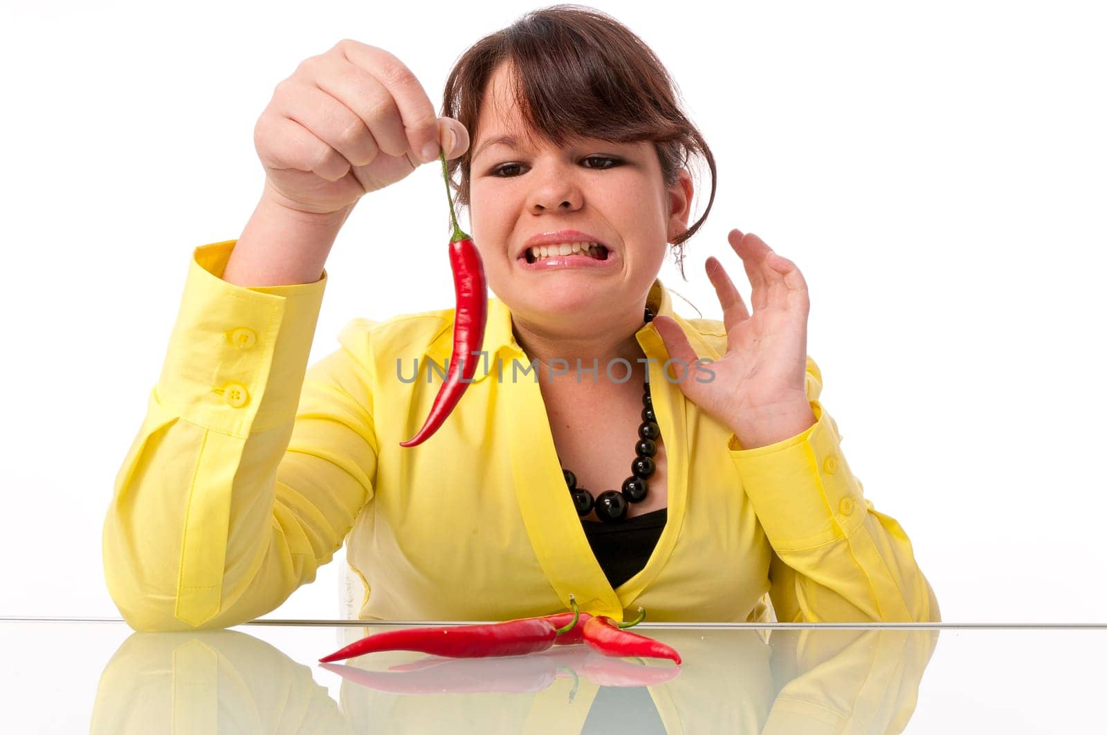 Teenage girl with long hair and yellow blouse is afraid of a hot chili pepper against a white background.