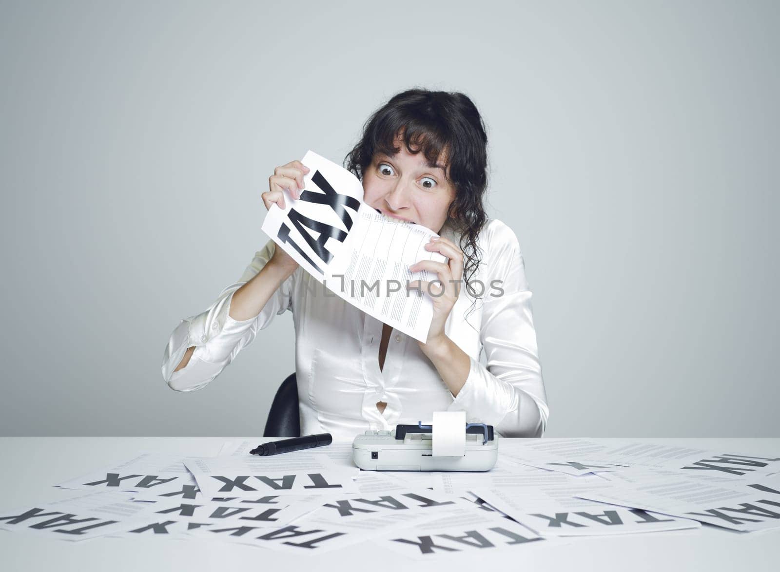 Young desperate woman at her paperwork covered desk biting a tax form