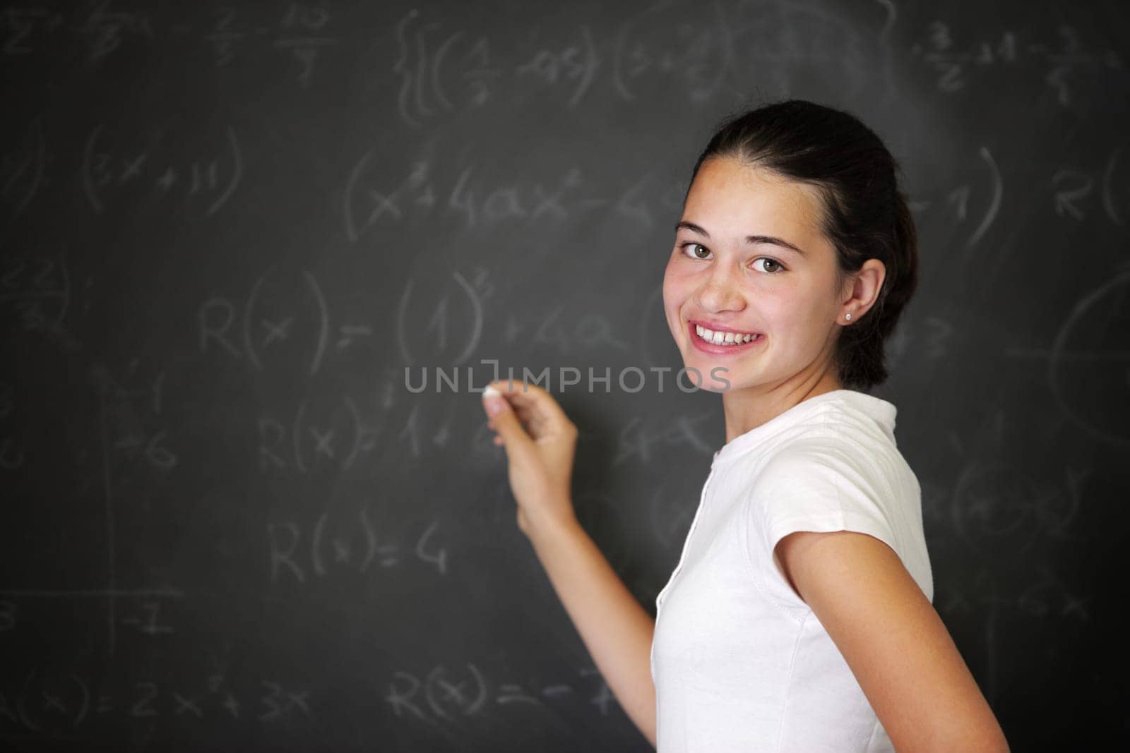 A young female student is standing in front of the blackboard going to draw something with a chalk.