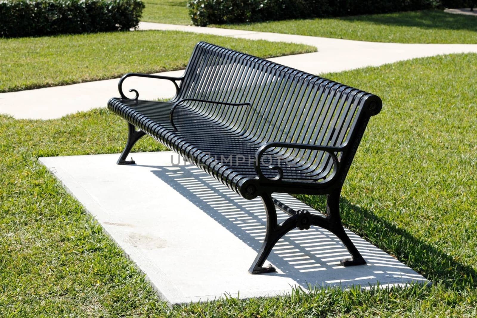 Black metal two seat park bench on a concrete slab surrounded by grass near a sidewalk and bushes on a bright winter day in Fort Lauderdale, Florida.