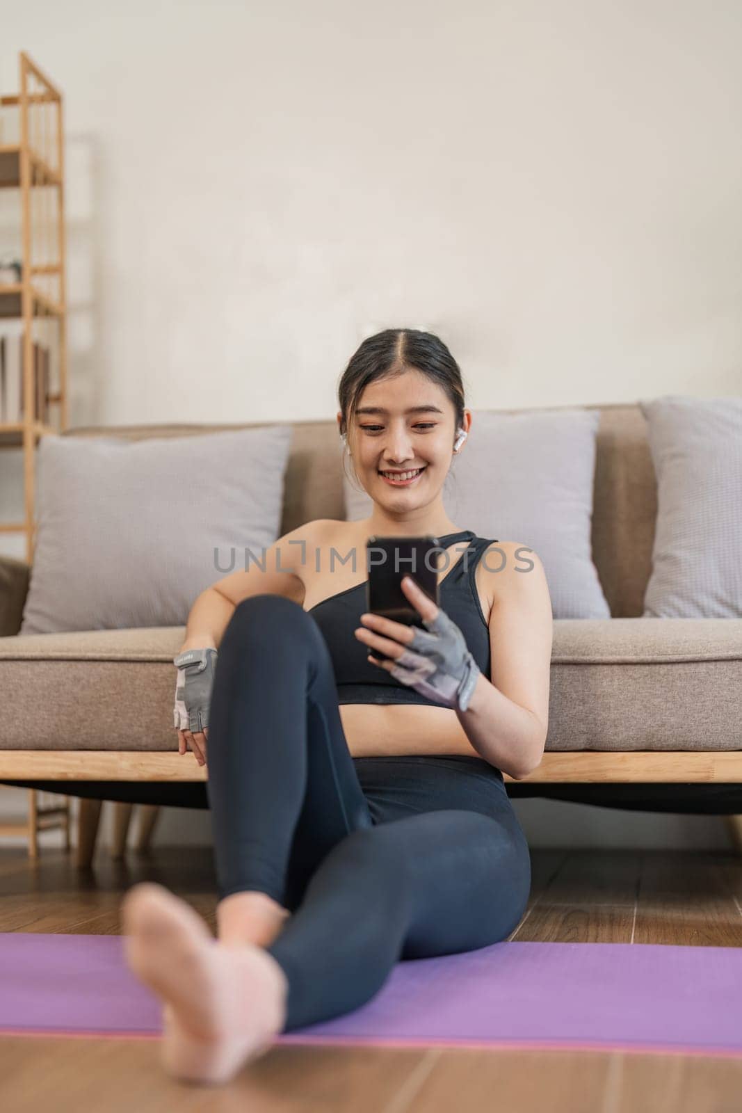 Lifestyle asian young fitness woman holding smartphone relaxing after workout at home. Smiling female using cell phone checking newsfeed on social media while exercise.