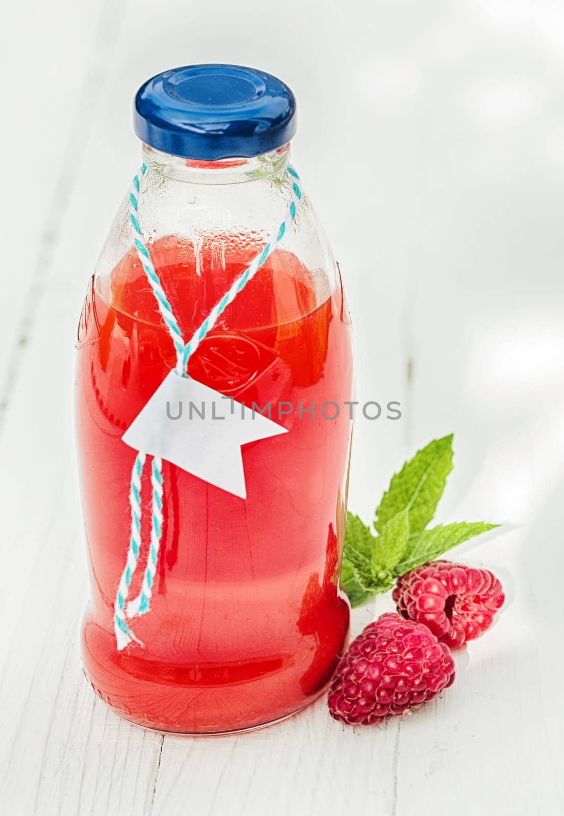 Homemade fresh raspberry juice in a glass bottle made from freshly squeezed fruit together with two fresh berries on a rustic white wooden background