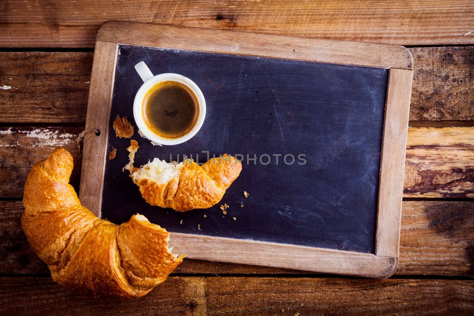 Overhead view of a fresh cup of coffee and a flaky croissant broken in two on an old school slate over a rustic wooden background, copyspace on the slate