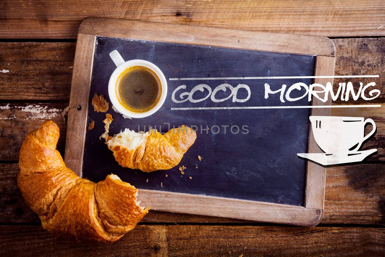 Good morning sign with a cup of fresh hot morning coffee and a broken croissant on an old school slate with a distressed wooden frame on a rustic table