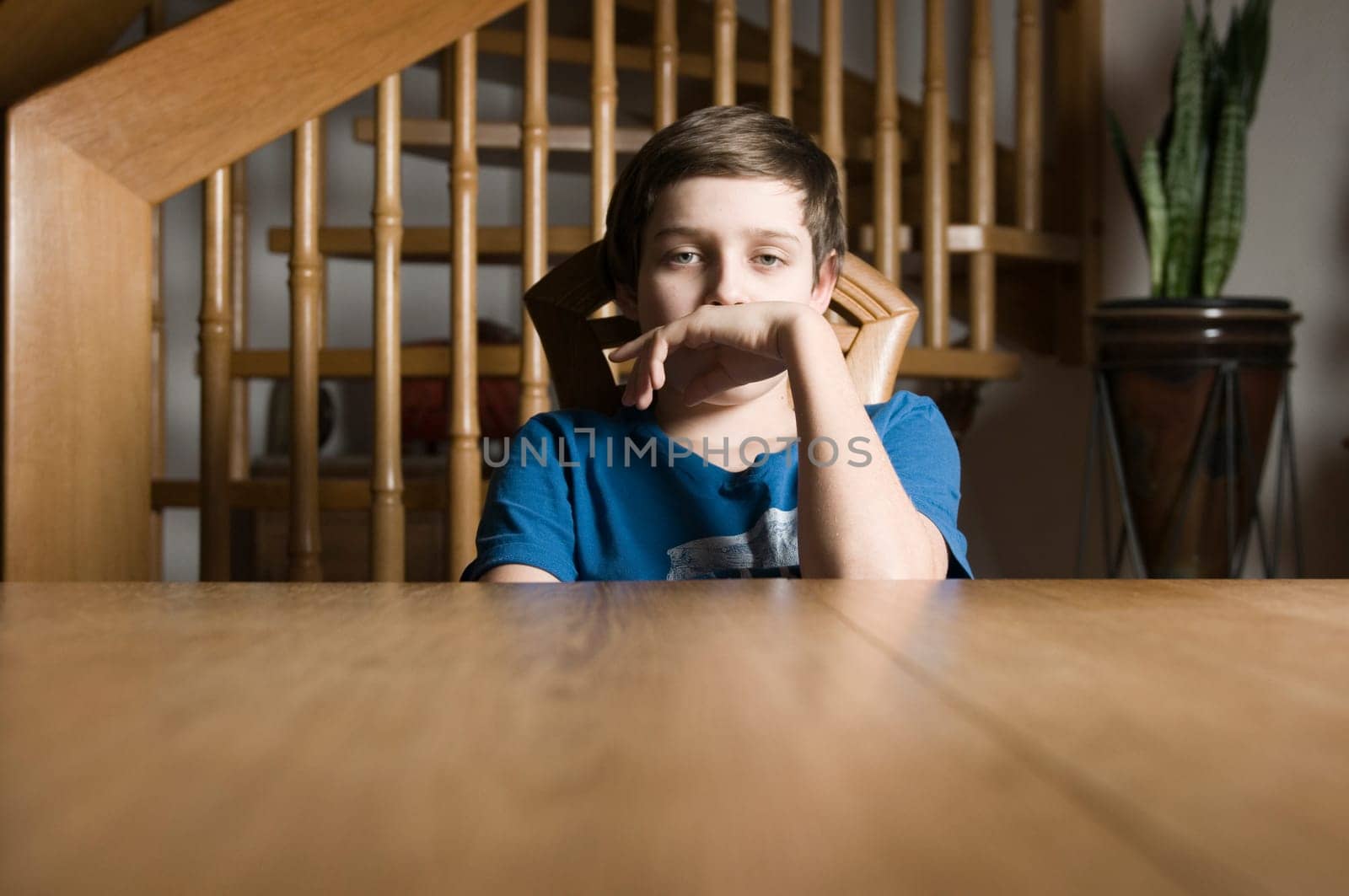 Frontal torso view of a young male teenager in a blue T-shirt sitting bored at a wooden table and looking at the camera with his hand held to his mouth