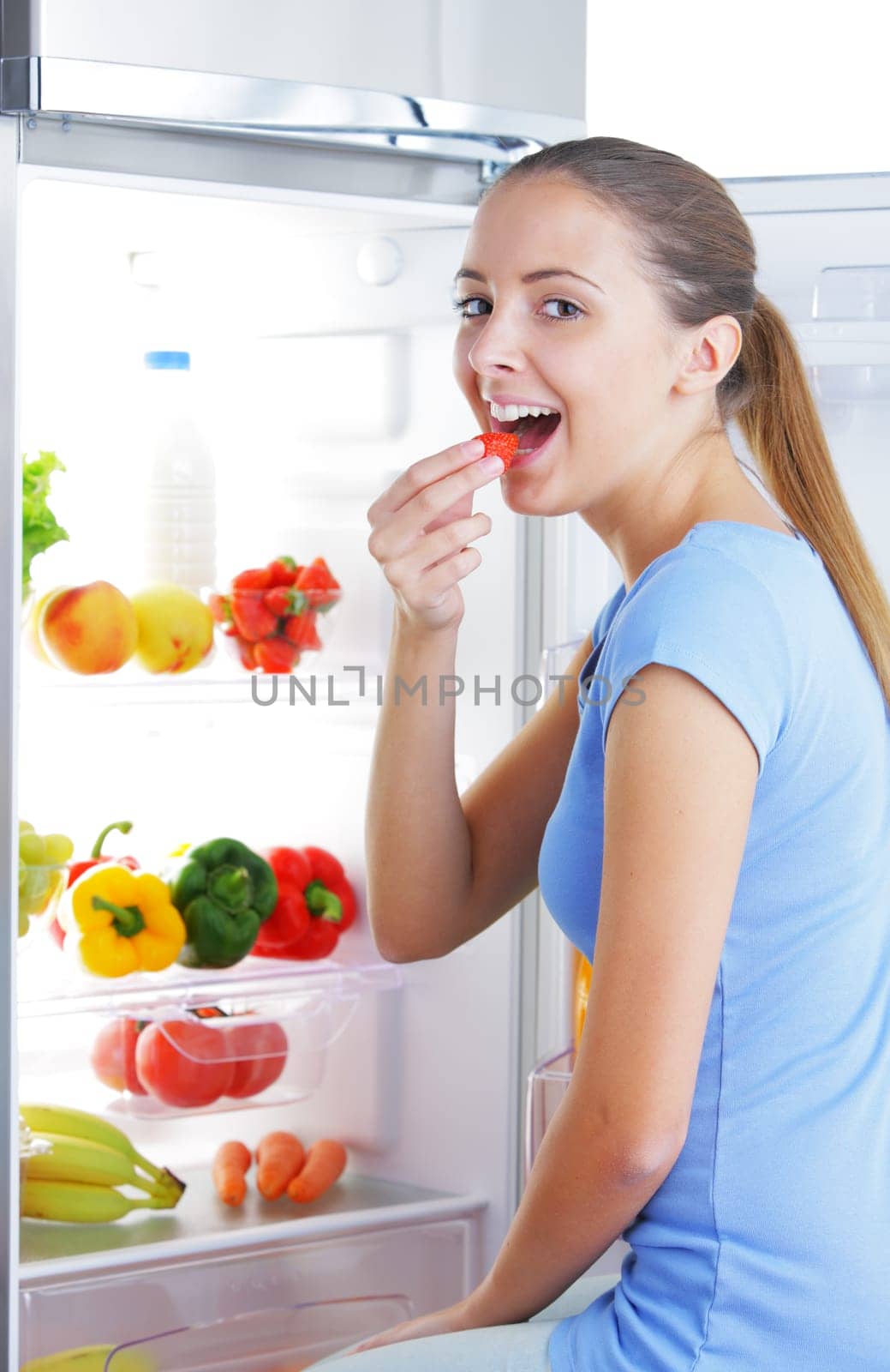 Smiling girl eating a strawberry near the refrigerator