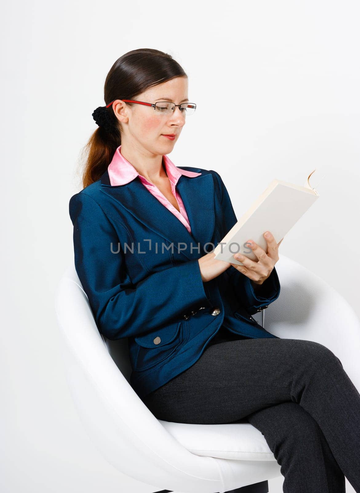 Young woman in a suit and glasses reading a book while sitting in a chair