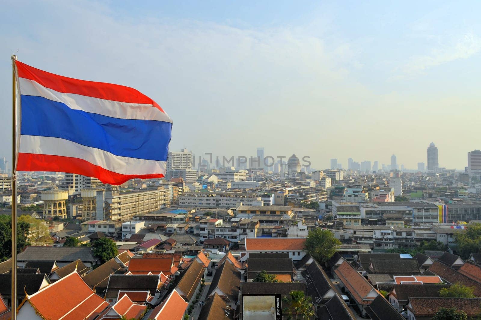 Skyline of Bangkok city by day with a Thai flag floating in the wind.