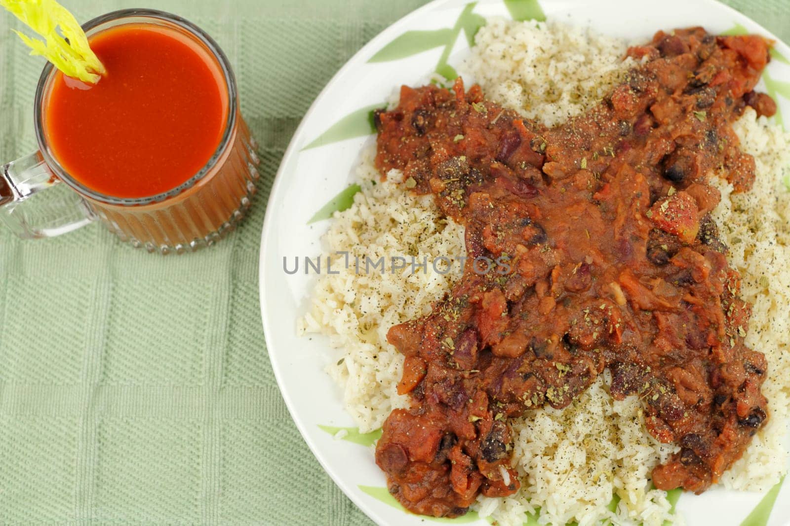 White dinner plate of brown rice topped with organic black, pinto and red kidney beans, diced tomato, oregano, and pepper. Drink is a clear mug of vegetable juice with a celery heart stalk garnish..