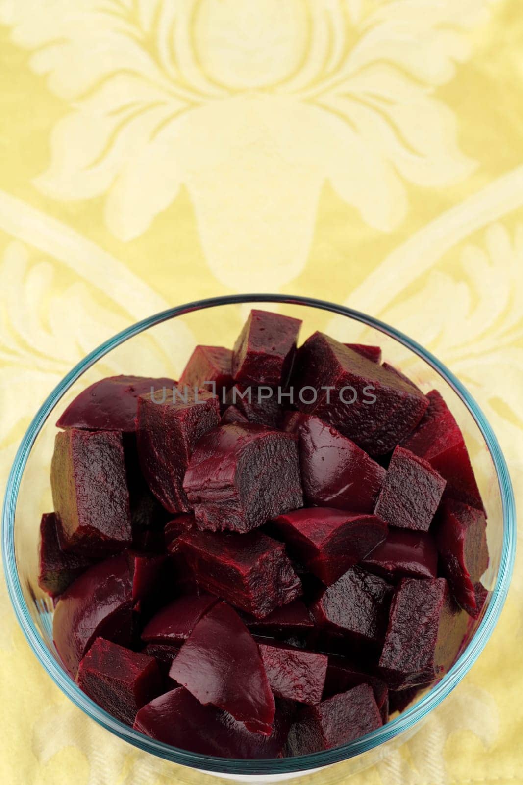 Red purple beets boiled, peeled, in chunks close-up in a clear glass bowl with a floral yellow gold place mat background..