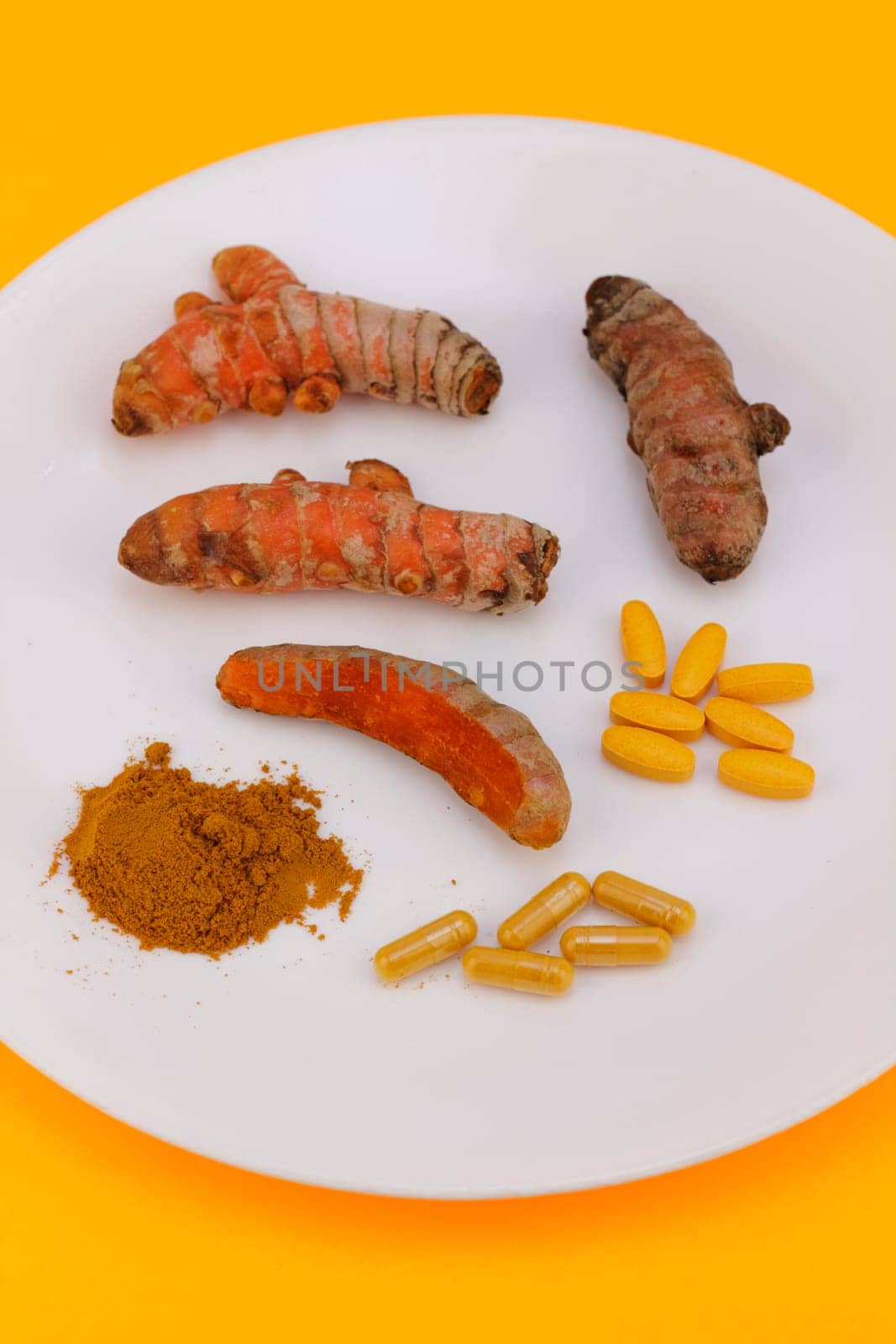 Turmeric displayed in ground powder, capsules and pills, whole raw roots or rhizomes peeled and partially peeled on a round white plate with an orange background.