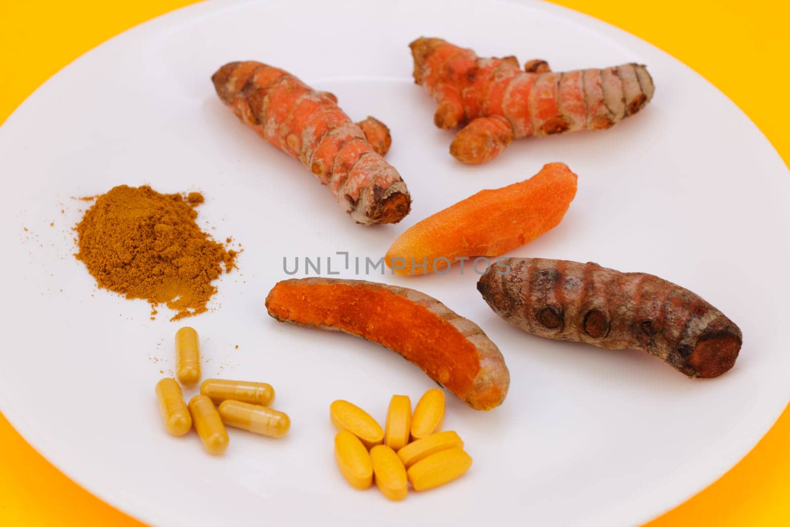 Turmeric ground into powder, in capsule and pill form, and the whole raw root or rhizome peeled, unpeeled, partially peeled, on a round white plate with an orange background.