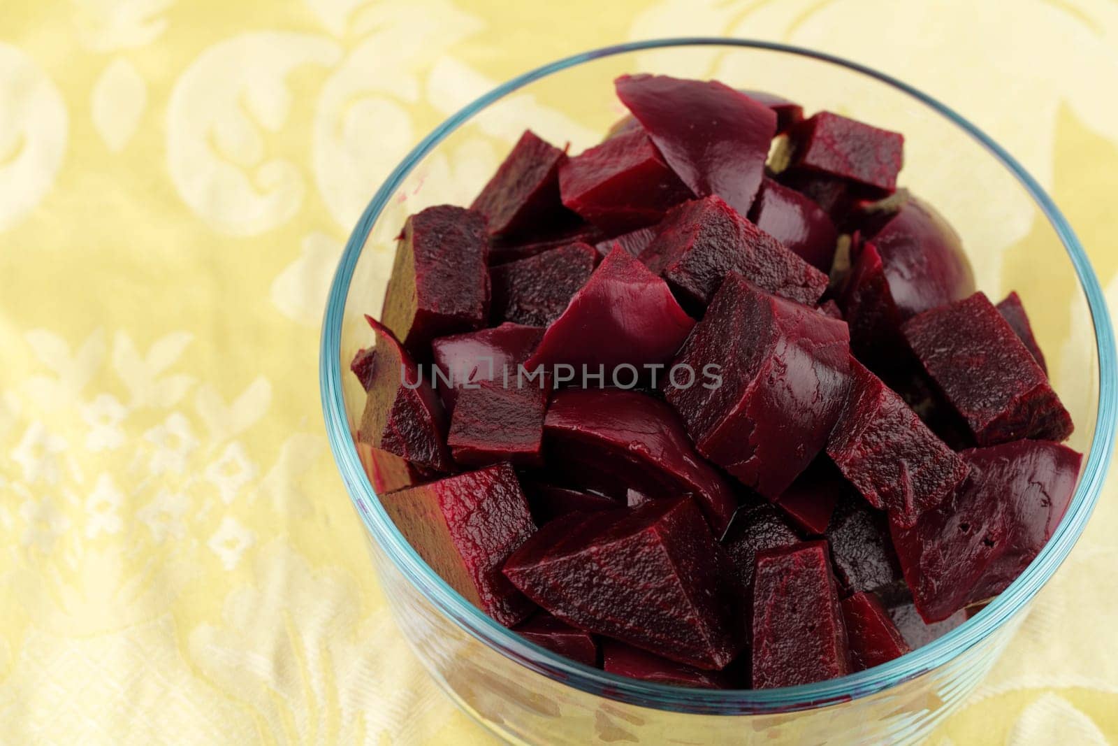 Dark red beets cooked, peeled and in chunks in a clear glass bowl set on a flowery  yellow place mat..