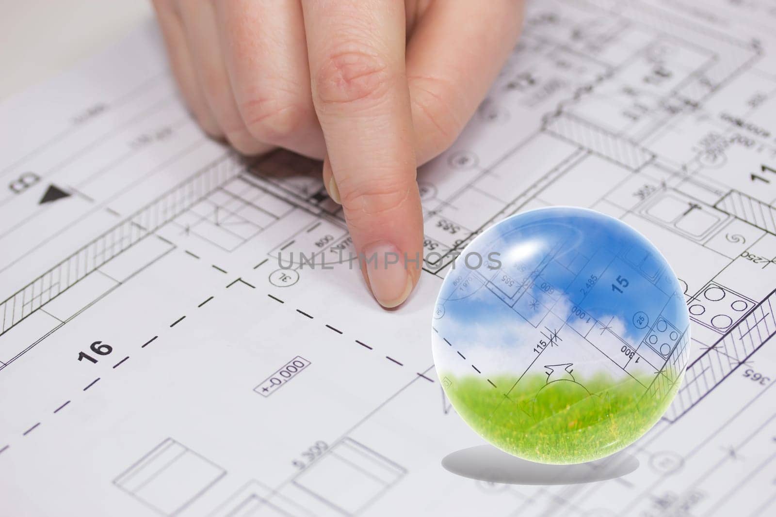 Finger from an unknown person is showing at the plan of the house. Blue sky, blueprint and green grass are in the glass ball.