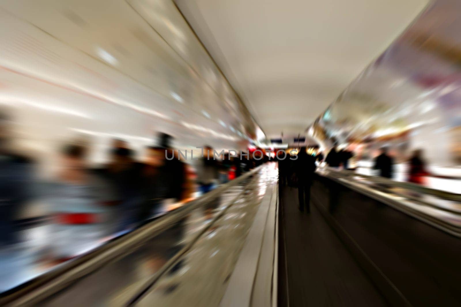 The railing of an escalator in a subway tunnel with passing people photographed with long exposure.