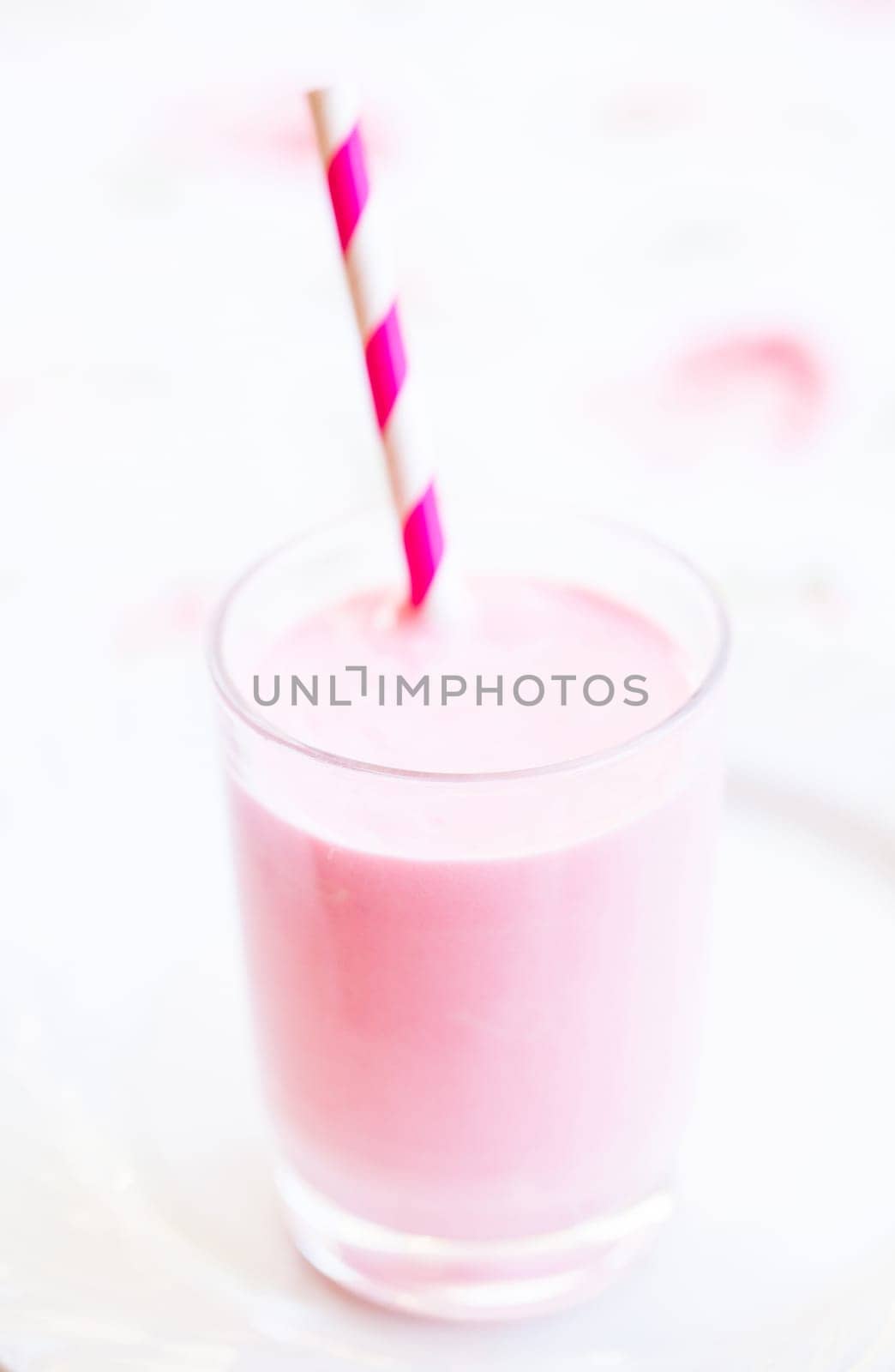 Closeup of raspberry smoothie made with milk and ice cream and with striped white and pink straw in glass on light background