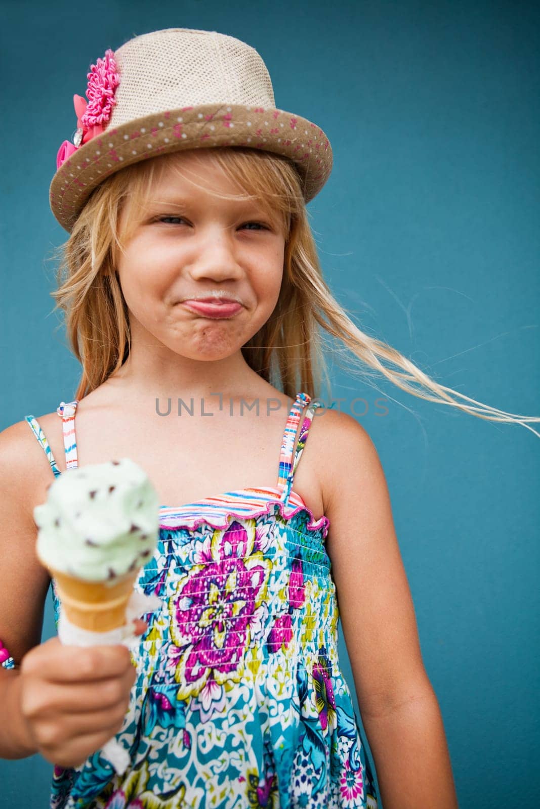 Cute young girl with funny expression holding ice cream cone outside against blue wall background