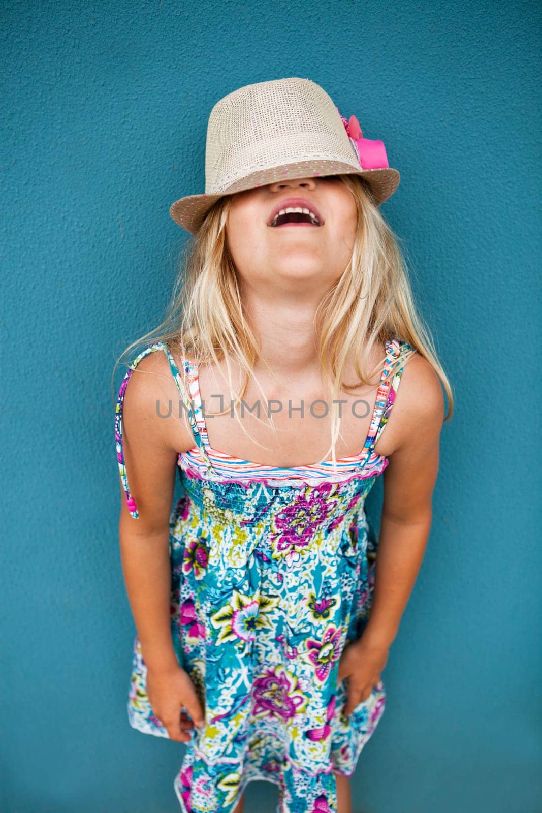 Portrait of stylish cute young girl against blue wall background with hat covering eyes and funny expression