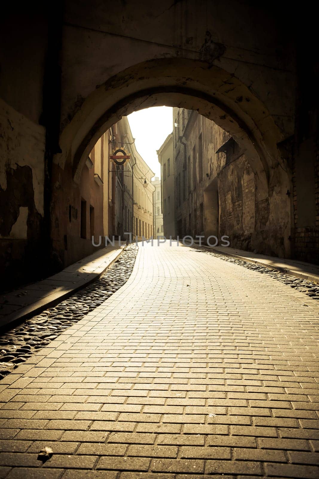 a fragment of the old town cobbled street in the sunlight in the down,empty street visible arch of the gate above the street.