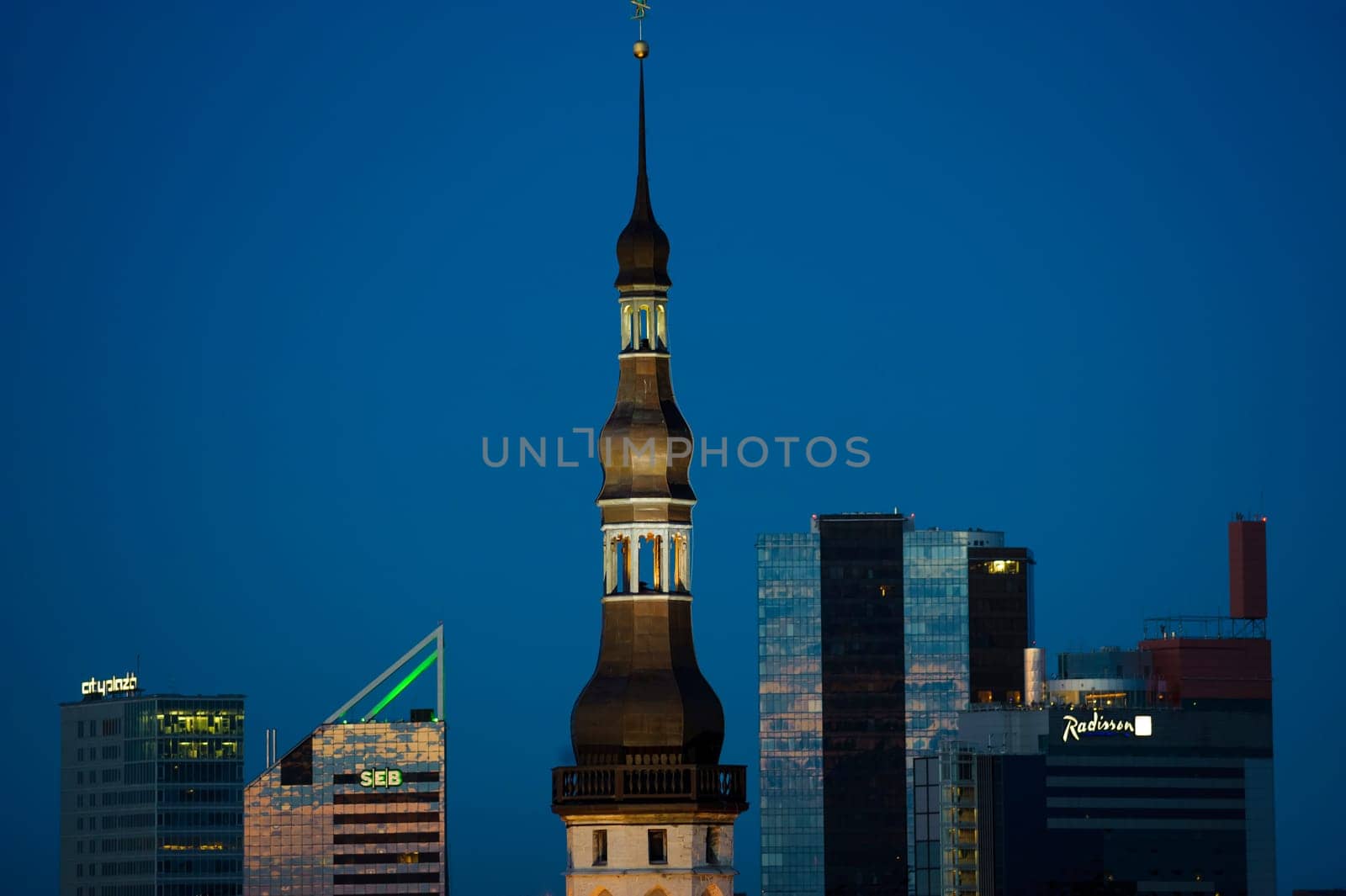 Photo presents fragment of the old town in Estonia, Talinn at night, visible illuminated church tower, modern office blocks in the background.
