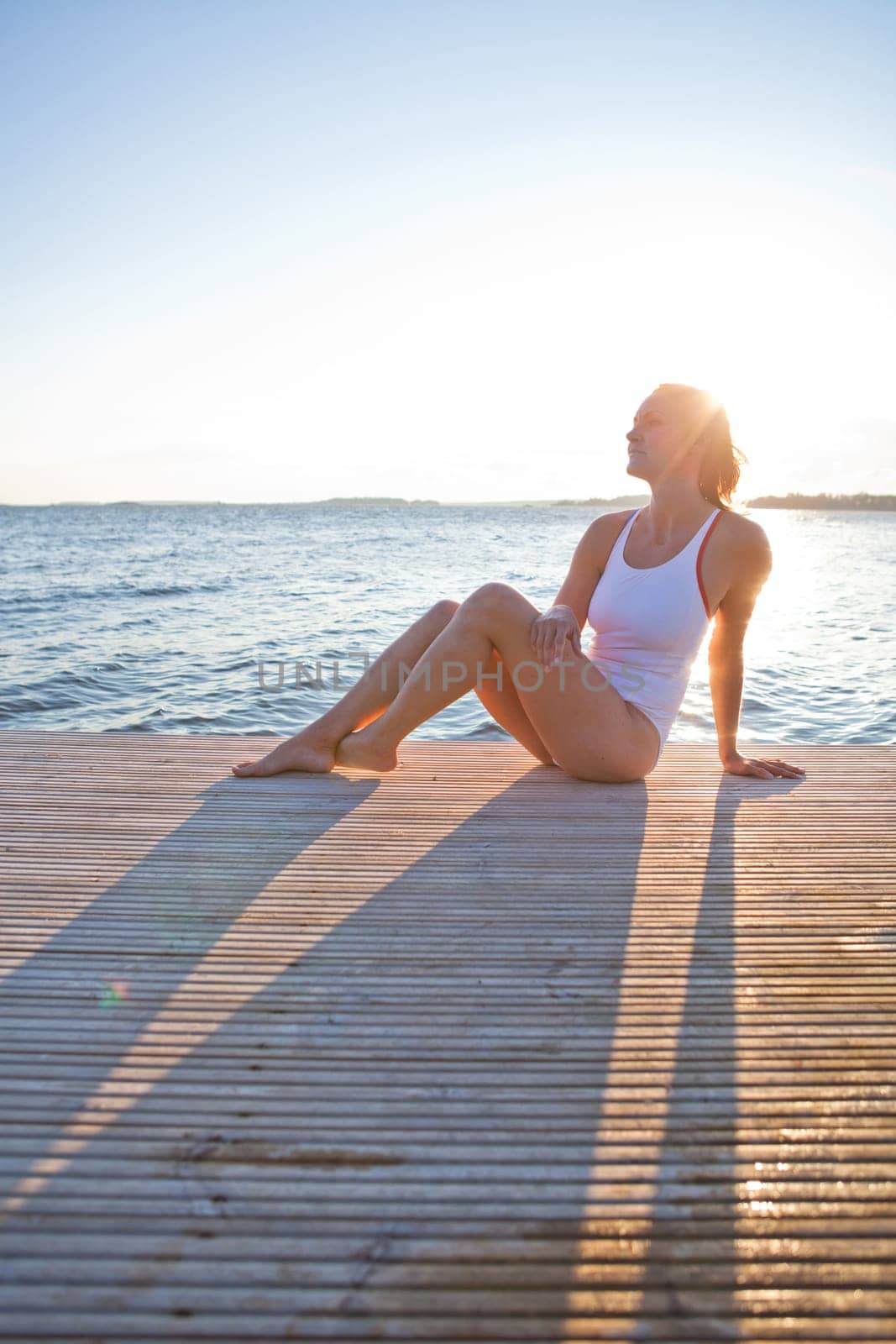 Attractive woman in white bathing suit sitting on wooden pier in nordic country during summer