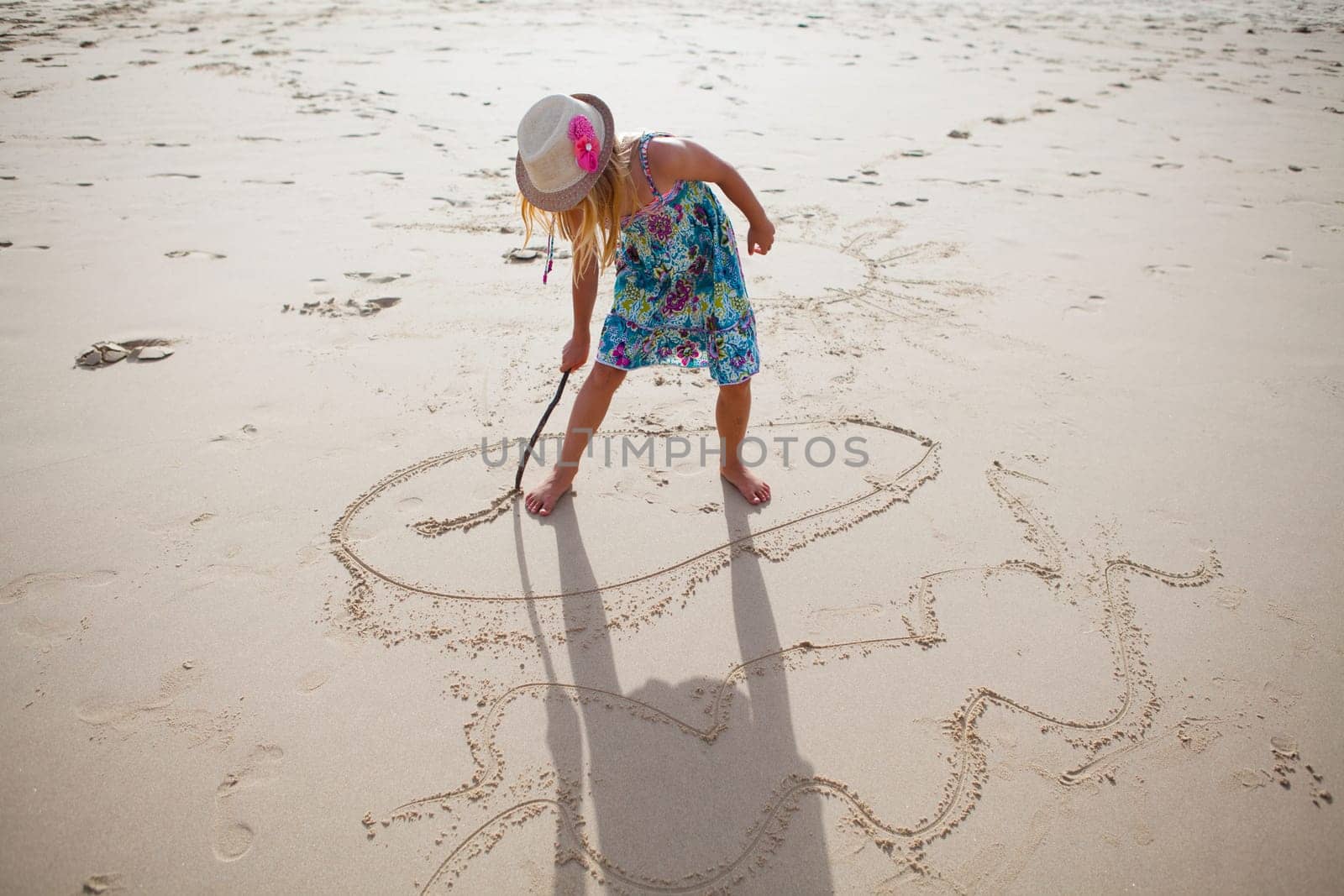 Young girl with summer dress and stylish hat drawing in sand with stick on beach