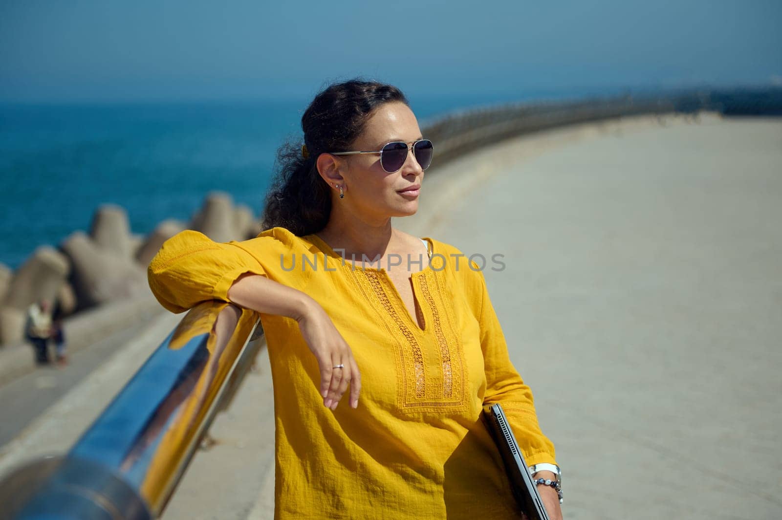 Confident portrait of charming Hispanic woman in sunglasses, holding laptop and looking into the distance, standing against the backdrop of the Atlantic Ocean on the city promenade. People. Lifestyle