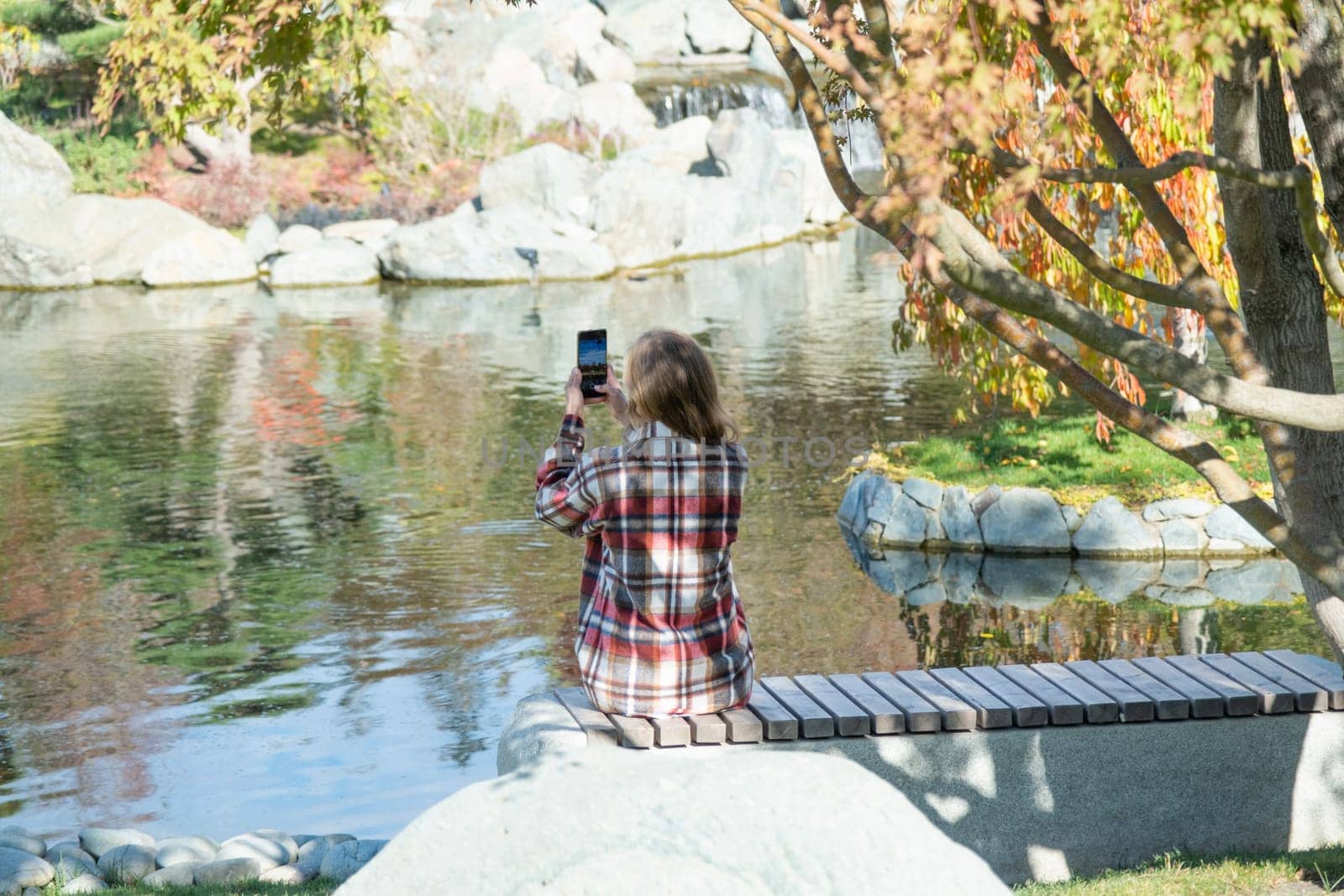 Woman in red plaid shirt enjoying nature standing in Japanese Garden taking photo on smartphone