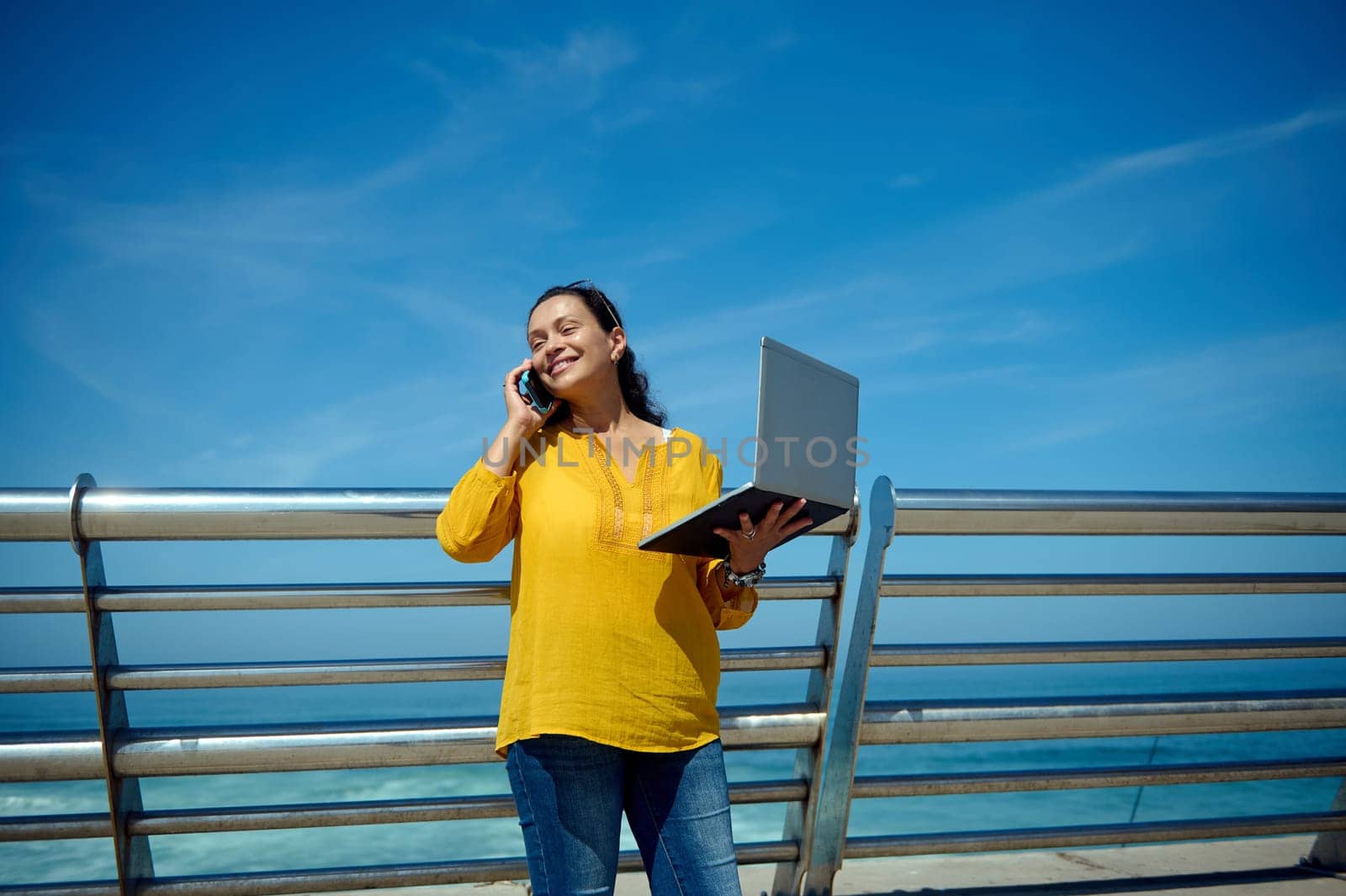 Waist up portrait of a middle aged confident successful freelance worker, manager, a charming business woman holding laptop and smiling broadly while talking on smart mobile phone outdoor. Copy space