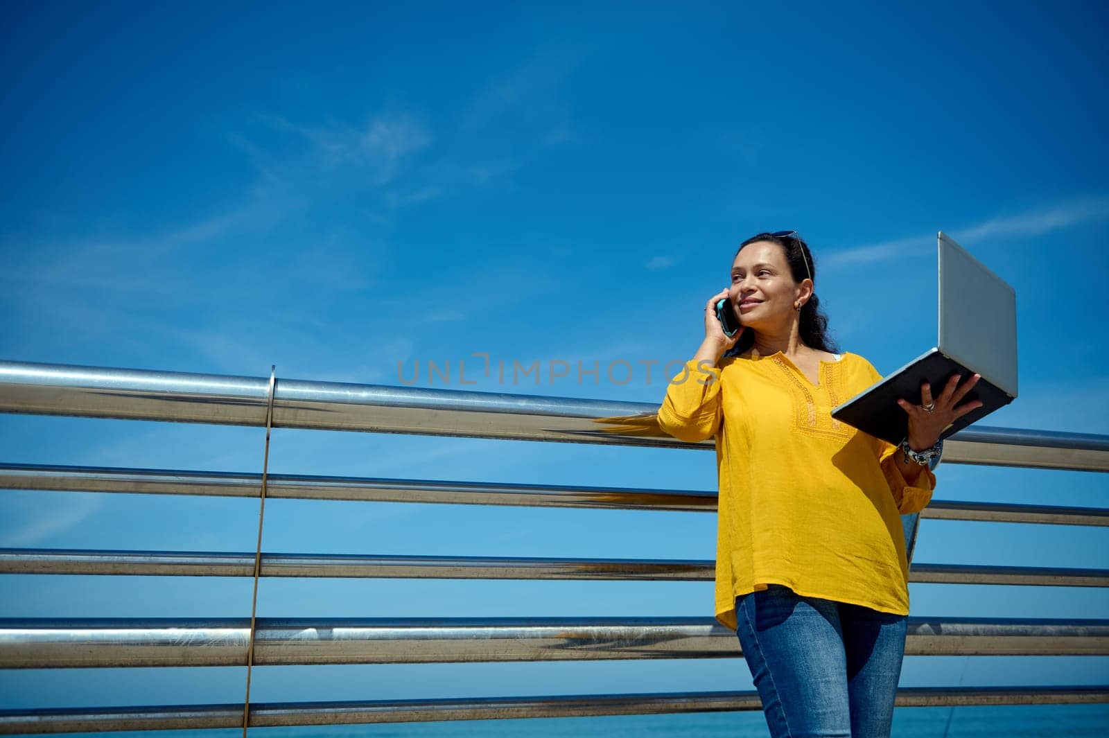 Authentic portrait of a confident successful businesswoman freelancer holding laptop and smiling talking on mobile phone, against a blue sky background with free advertising space for promotional text