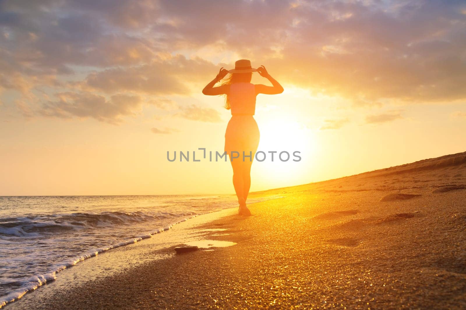 Female silhouette, rear view, with hands raised to the hat and hair flowing. Seascape during golden sunrise with beautiful sky, young happy woman walking along the seashore. A girl looks at a magical sunset.