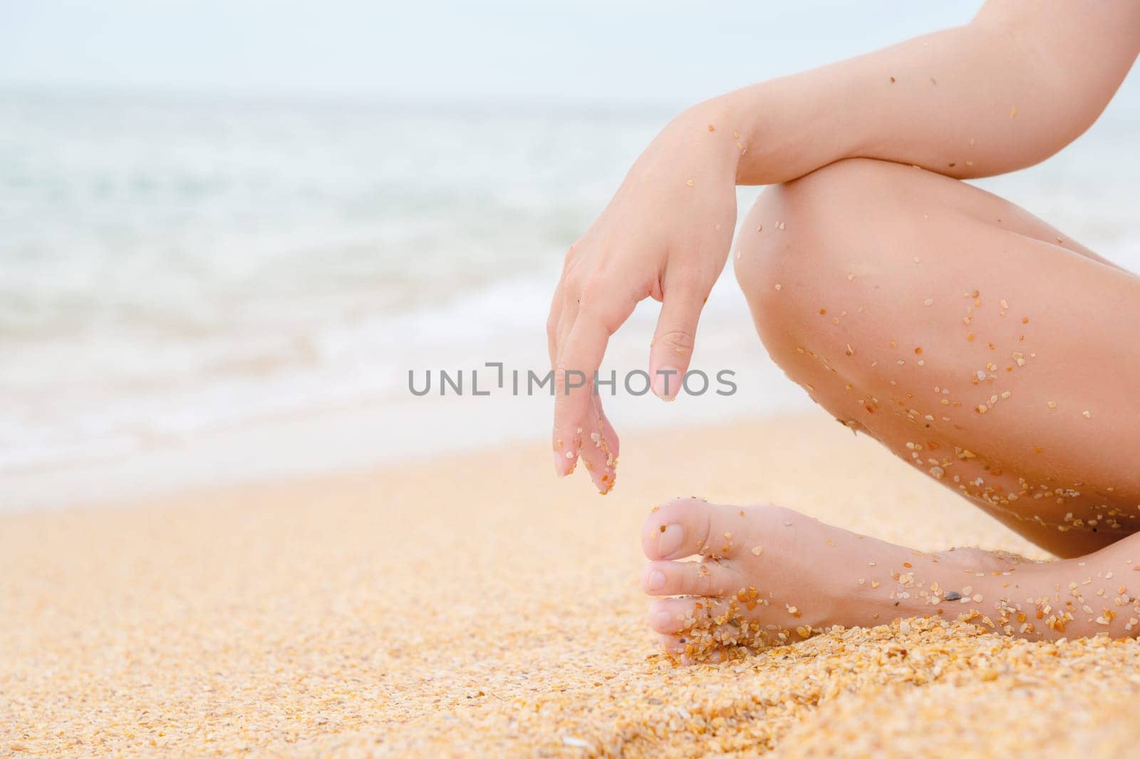 woman sits on the shore and relaxes. A woman's leg and hand on her knee, a girl sitting on the sea sand on the beach on a summer day, close-up by yanik88