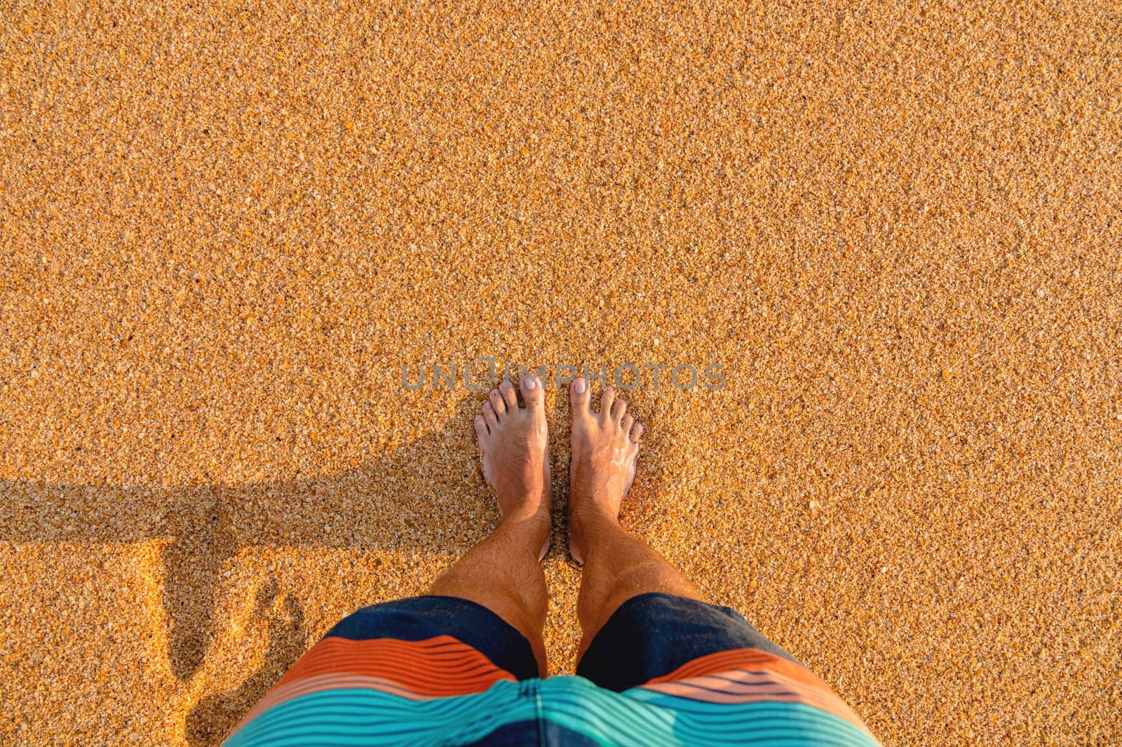Top view of male legs in colored shorts on golden sand in sunlight. First person view of sandy beach in summer on vacation by yanik88