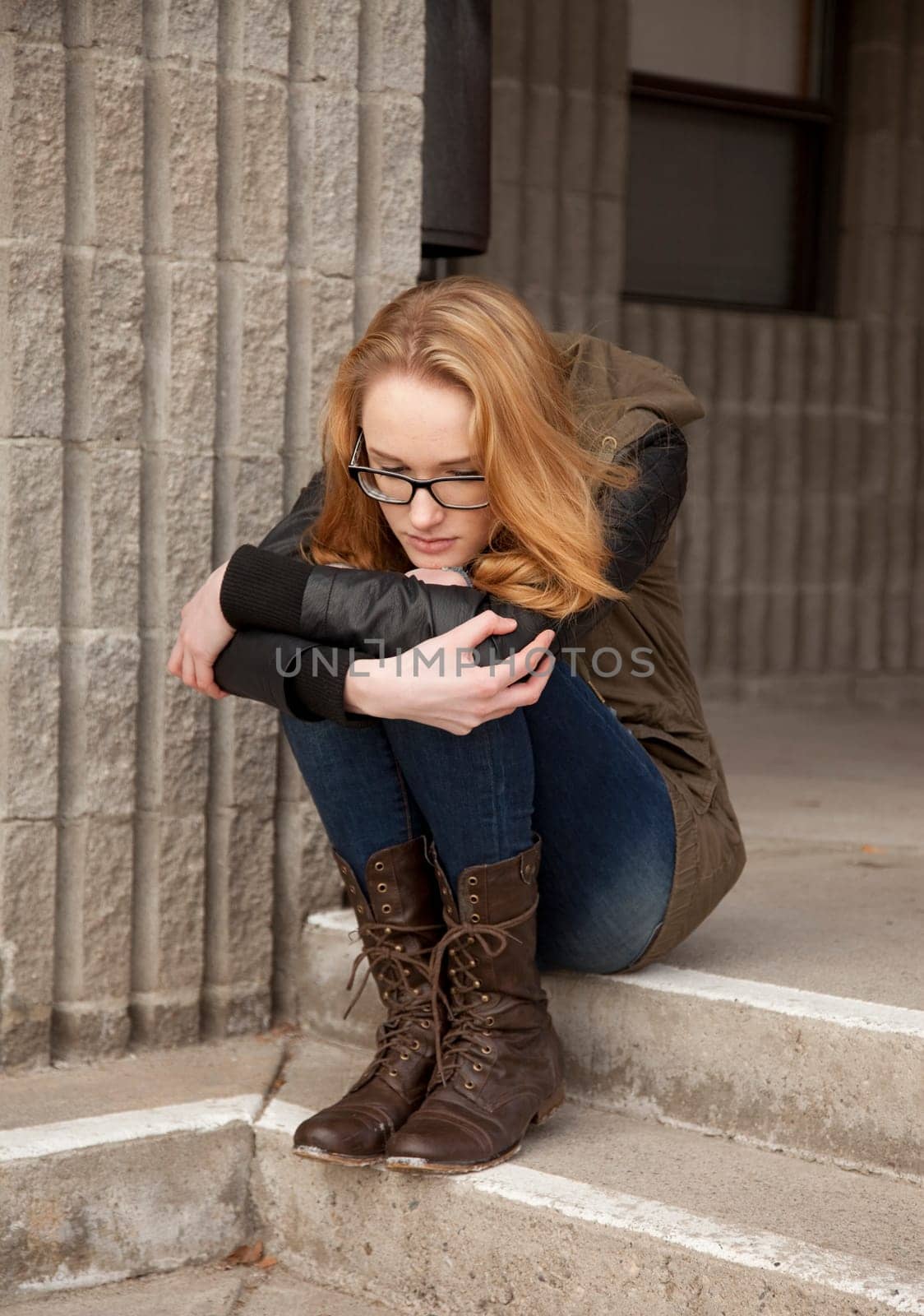 Outdoor photo of young teenage girl seated on concrete steps, hugging knees, distraught facial expression.