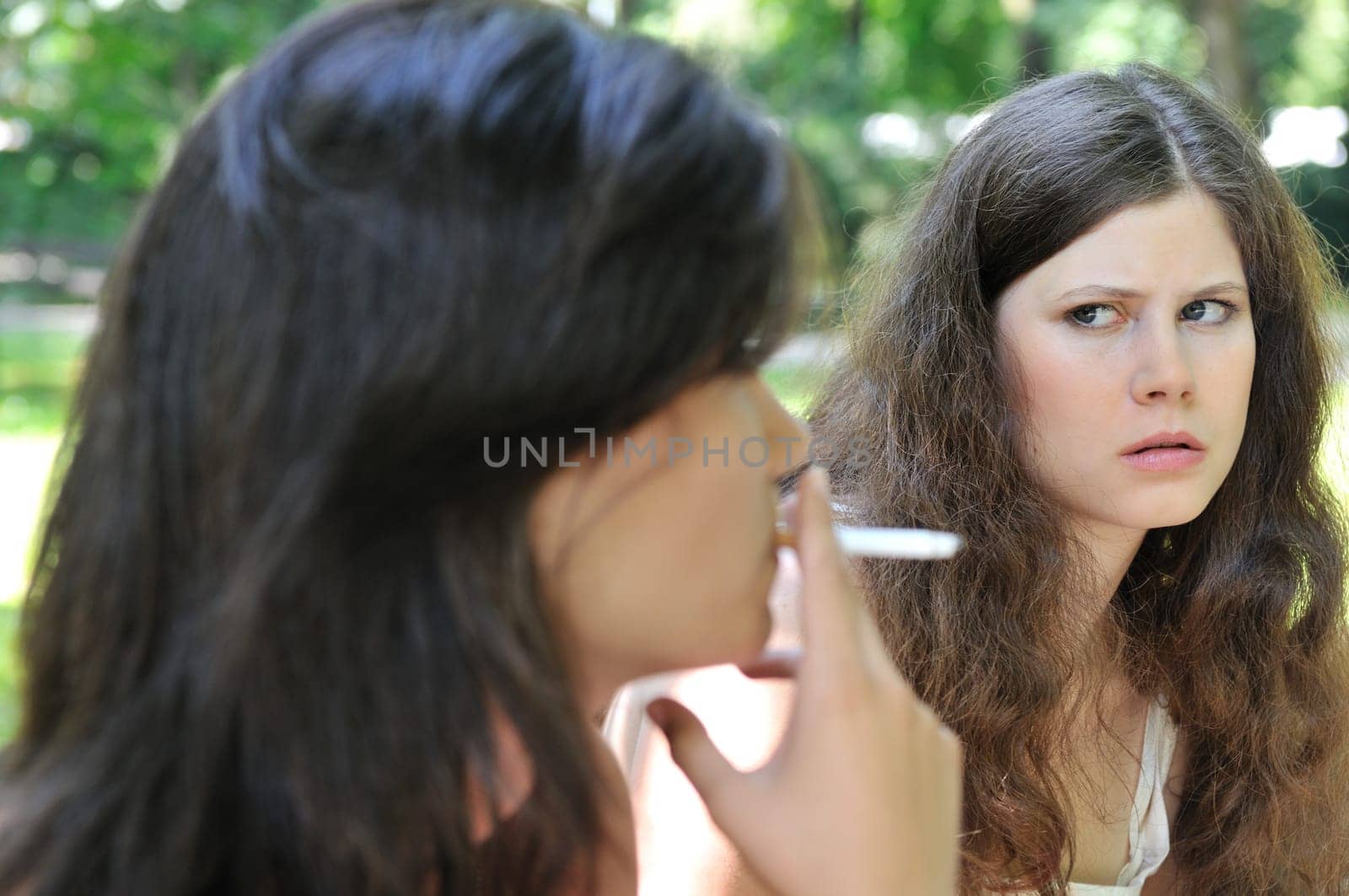 Youth culture - two young people outdoors, one woman smoking cigarette annoys and iritates another girl