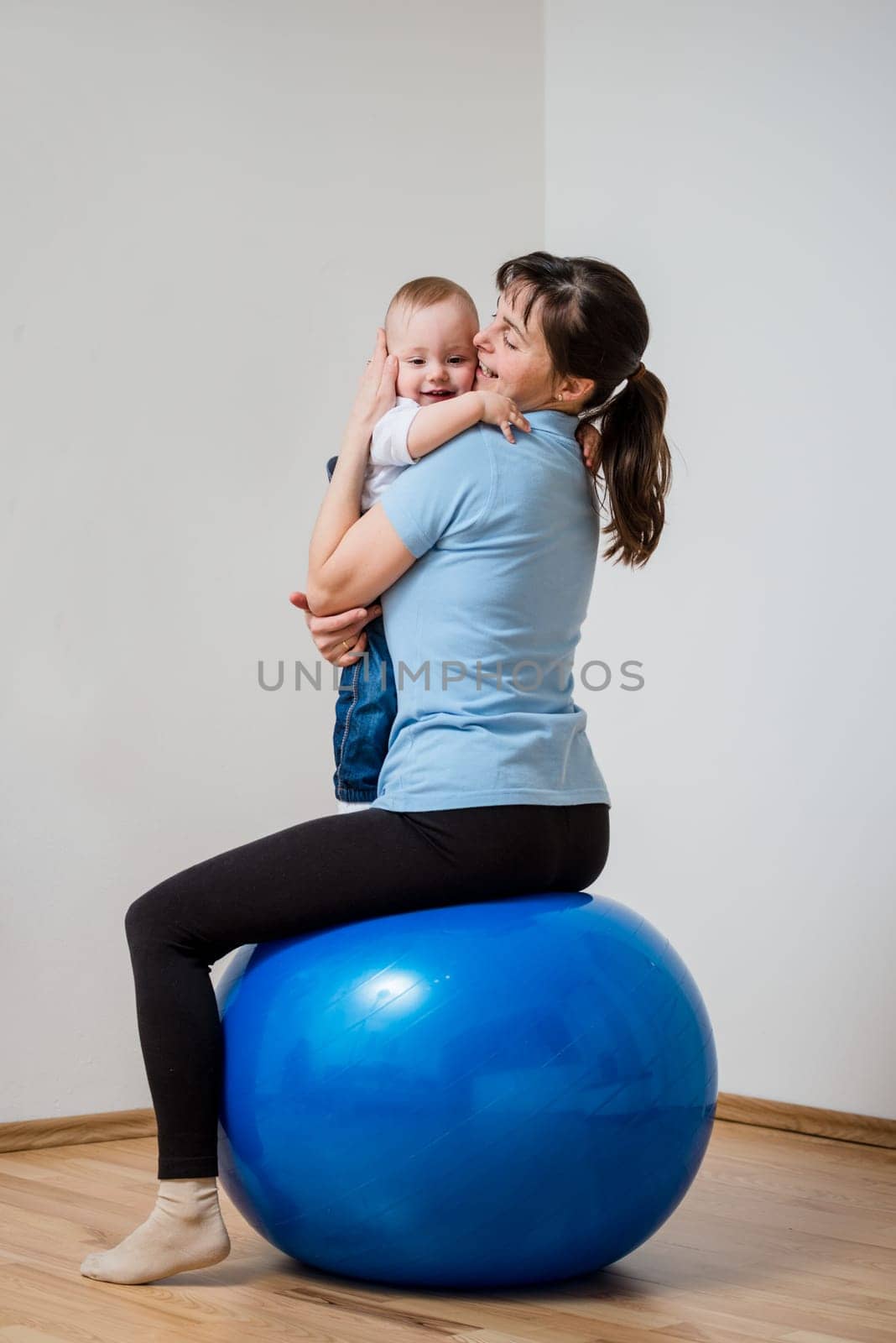 Mother exercises with blue fit ball at home holding her baby on hands