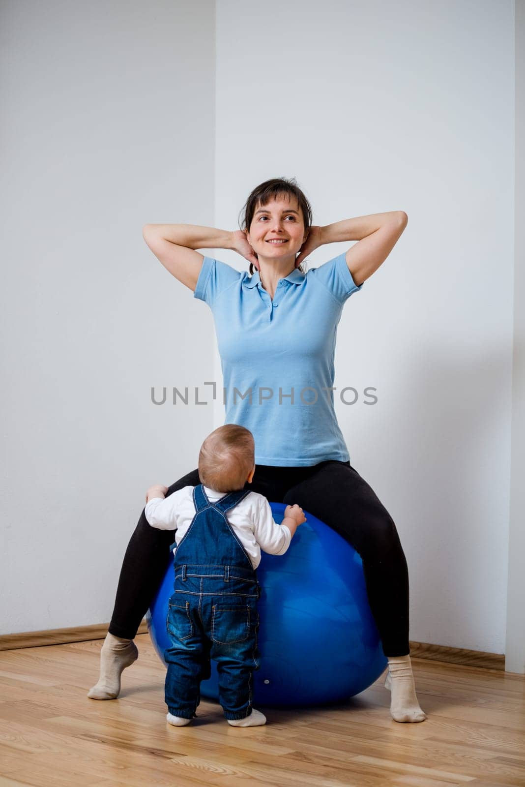 Mother exercises with big blue fit ball at home and her baby is watching her