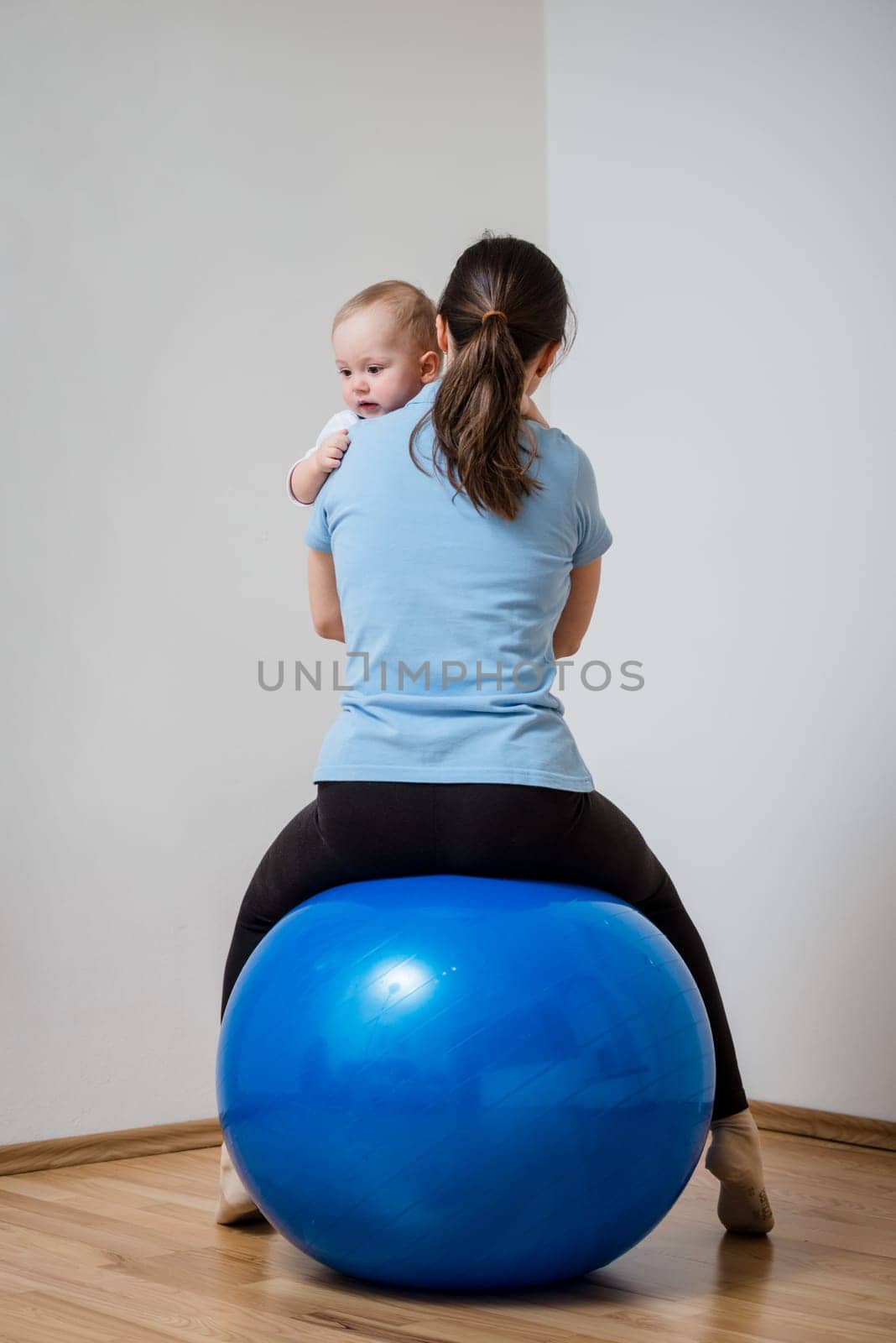 Mother exercises with blue fit ball at home holding her baby on hands