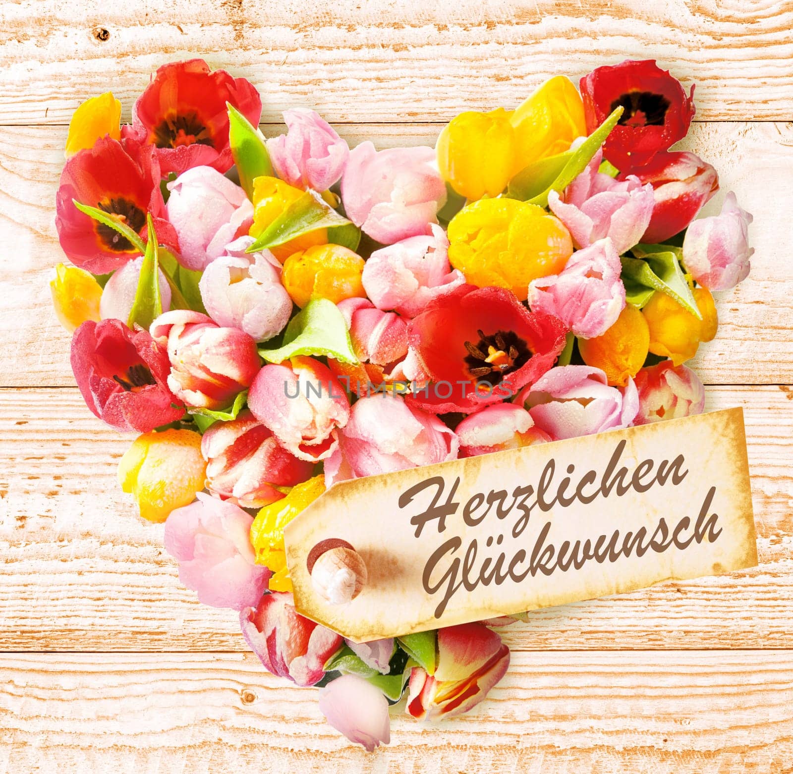German birthday wishes on a colourful floral heart formed of fresh spring tulips in vibrant colours arranged on a rustic wooden background with a tag saying - Happy Birthday or Herzlichen Gluckwunsch