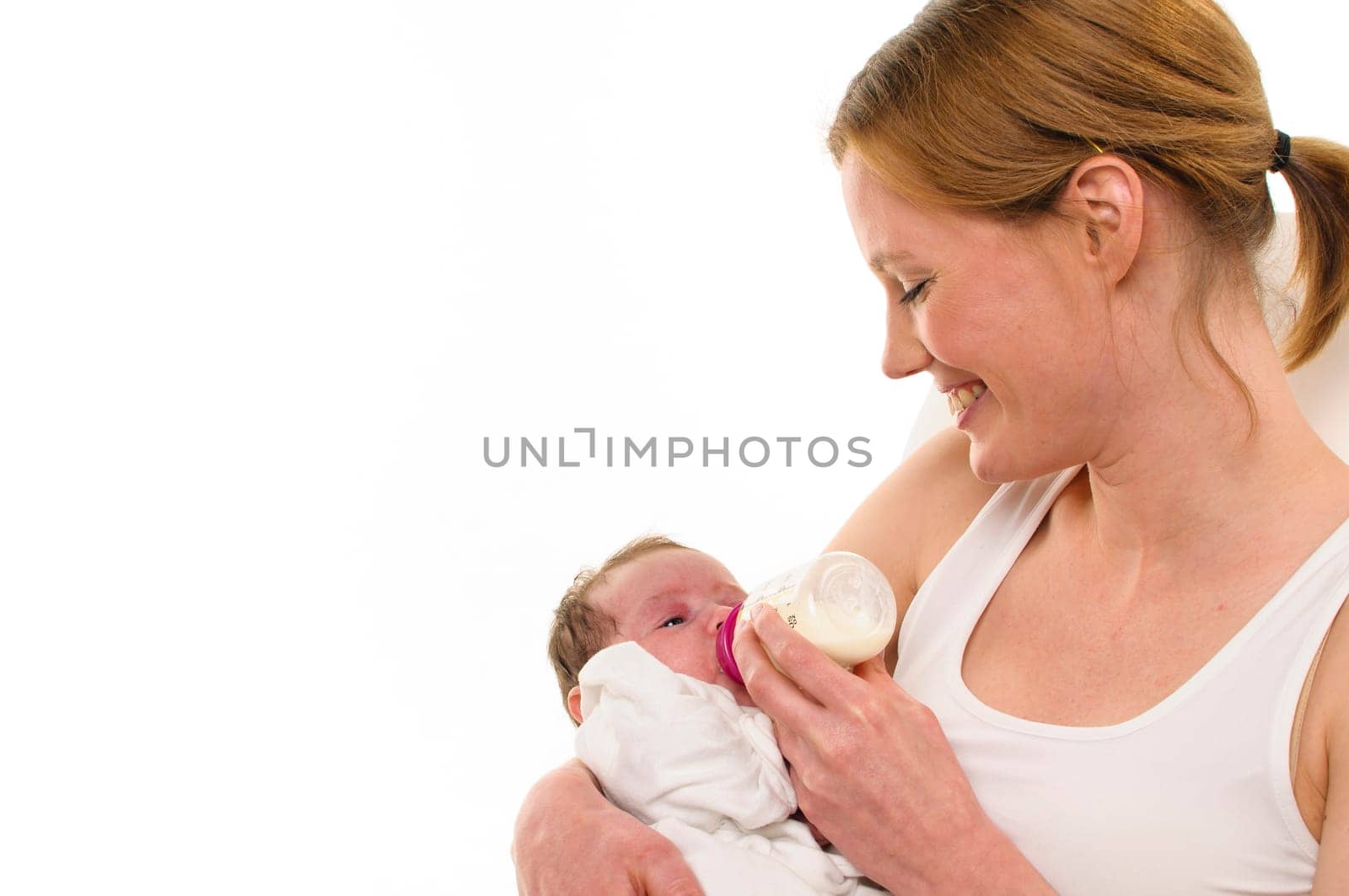 An adult woman with a white shirt tenderly holds an infant,also dressed in white,in her arms and gives him his bottle,exposed against a white background.