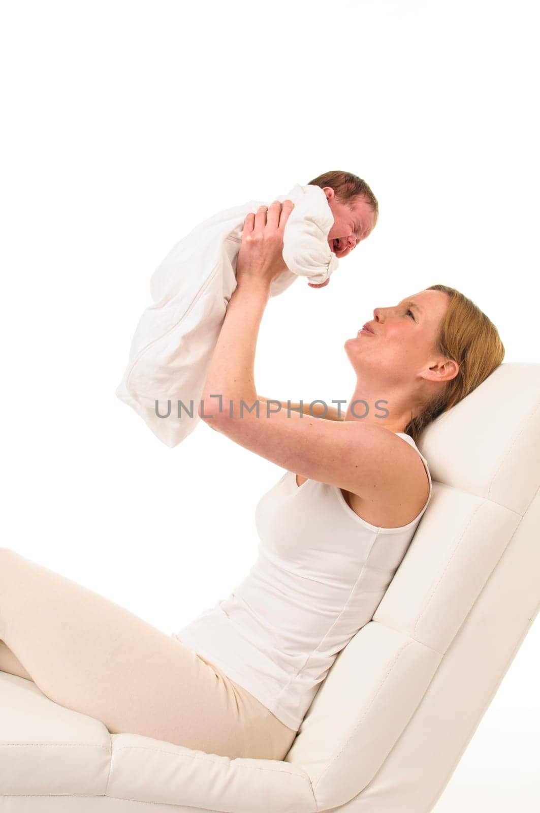 An adult woman with a white shirt lies on a white couch and holds an infant dressed in white in front of her in the air,exposed against a white background.