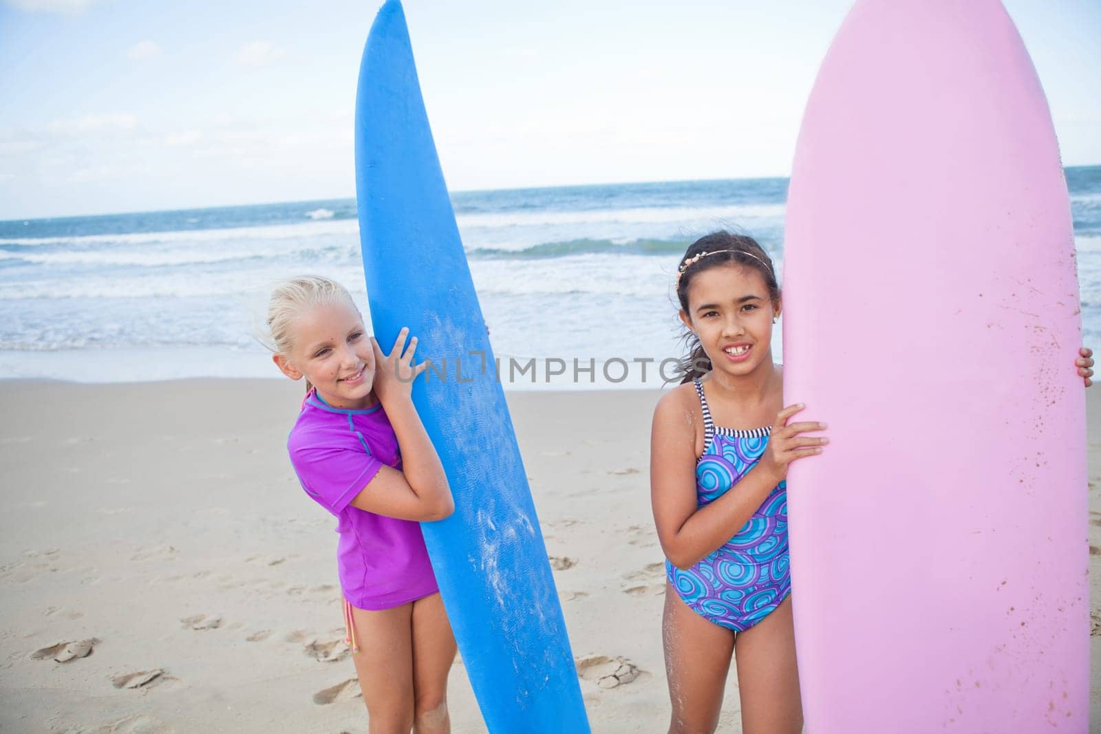 Two cute smiling young girls holding surfboards upright at the beach with ocean in background